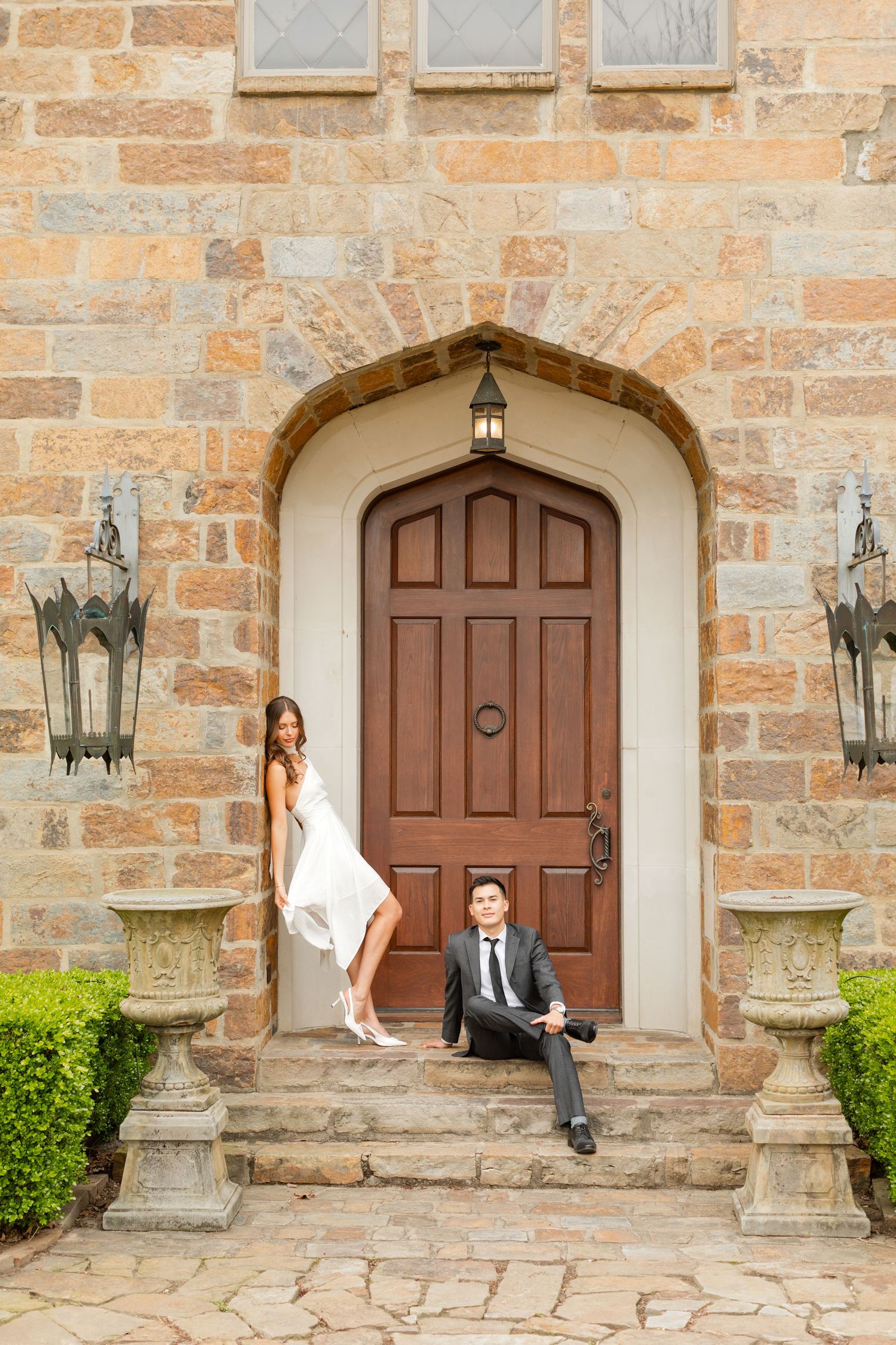 A couple poses playfully in front of a stone building with wooden doors and vintage lanterns. Goodwin Manor.