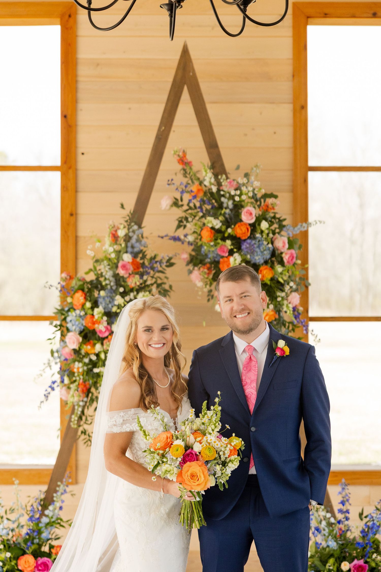 Wedding couple poses in front of colorful floral arch with bright summer blooms.