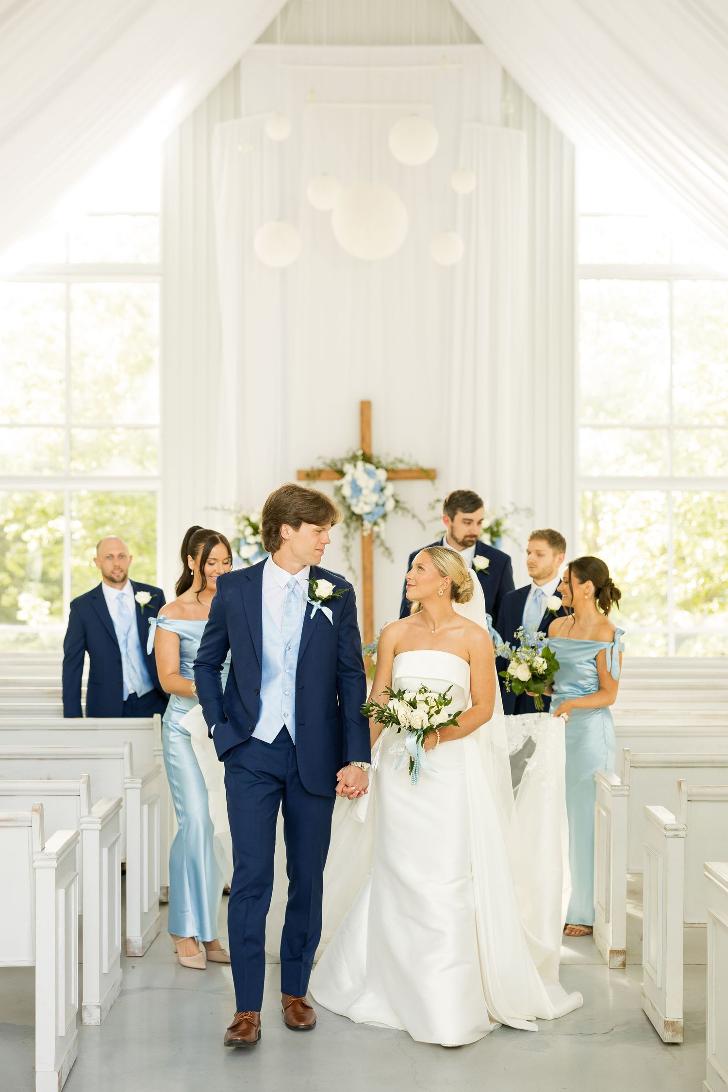 Wedding party in navy suits and light blue dresses posing in a bright white chapel.Cold Springs Events