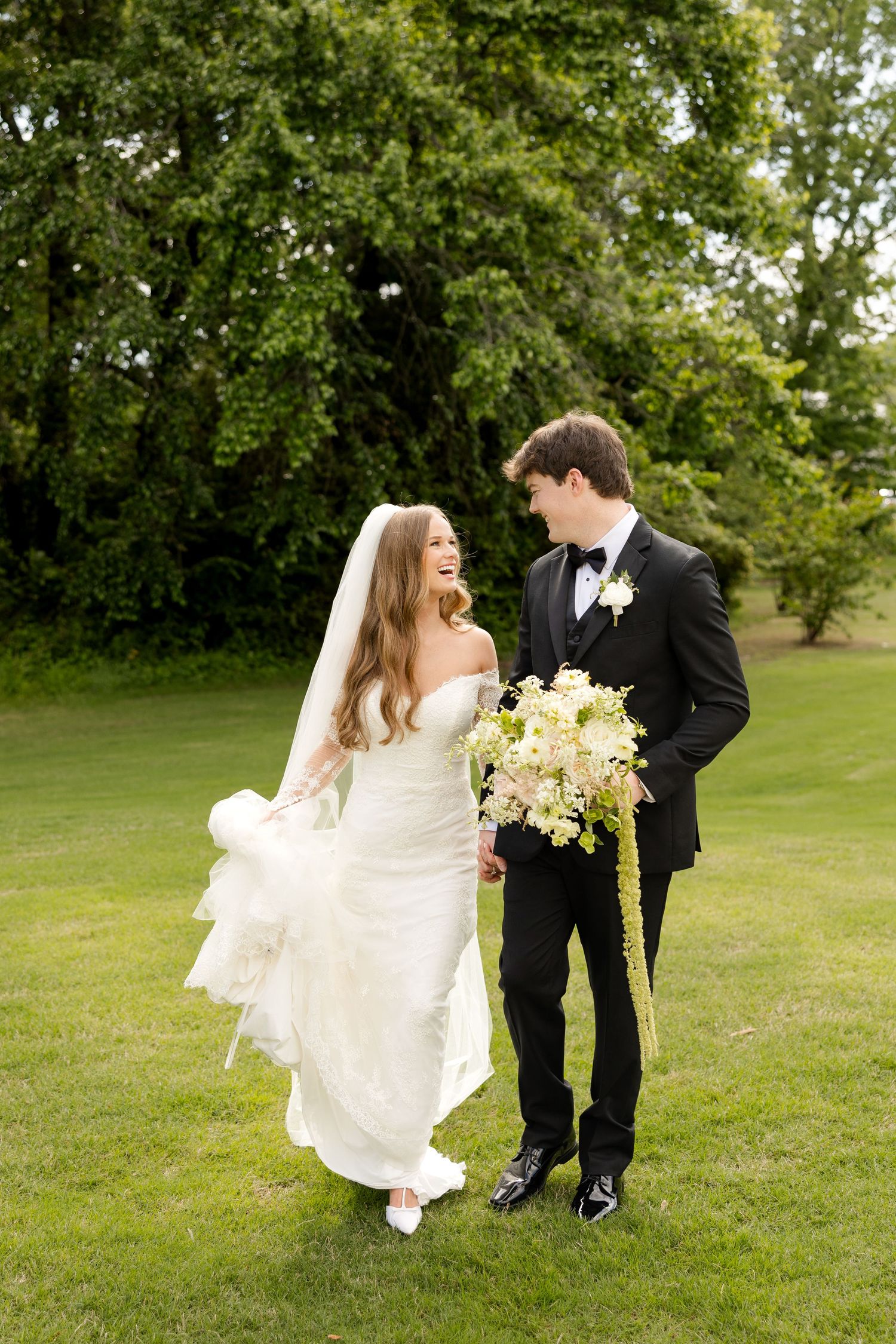 Bride and groom walk hand-in-hand on green lawn, she lifts white gown while holding bouquet, both smiling joyfully.