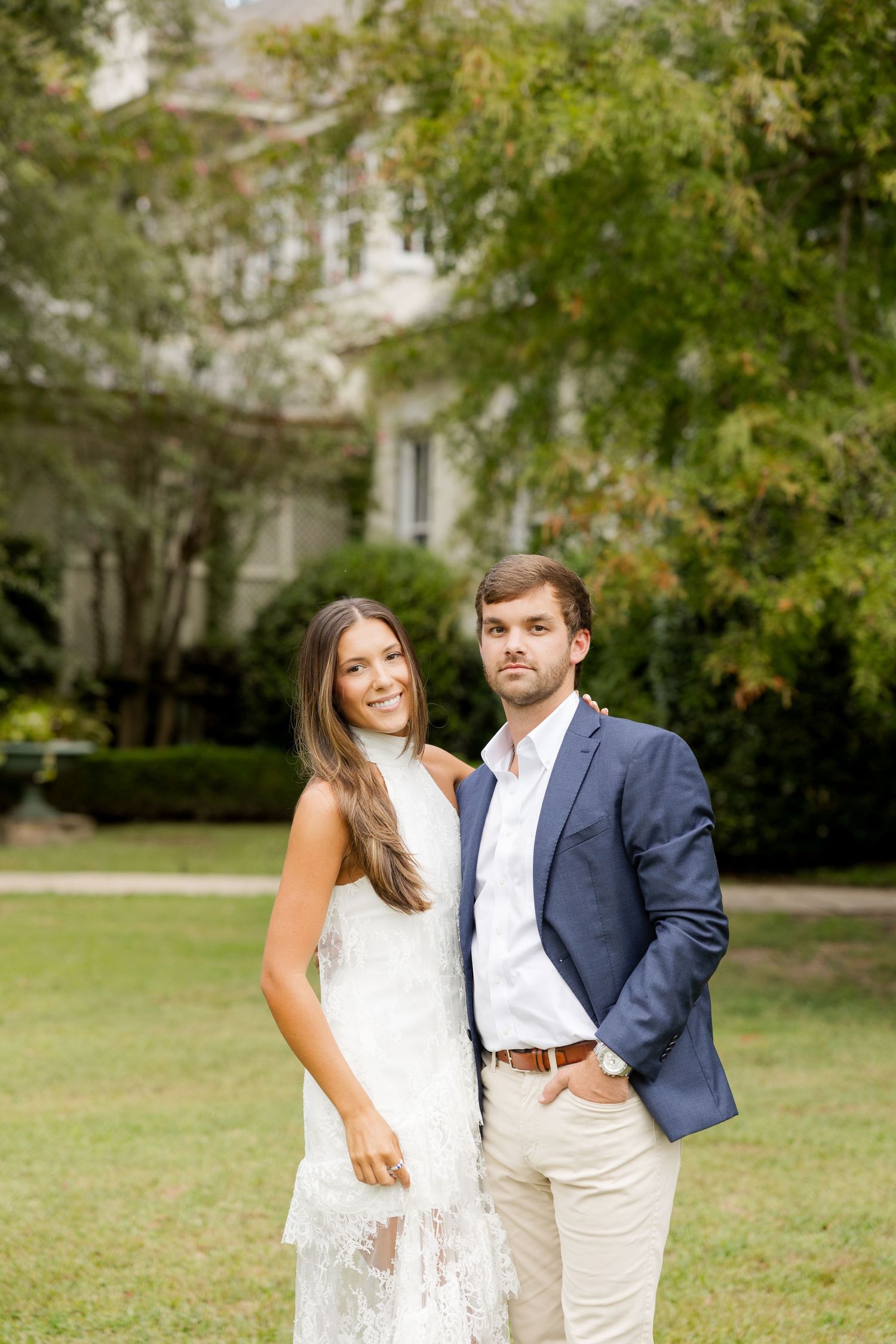 A couple poses together on a lawn in front of a stately white mansion on a sunny day.