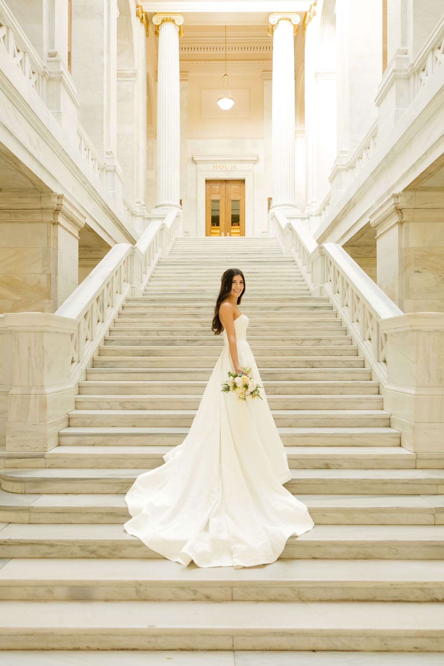Bride in white gown stands on ornate marble staircase with bouquet in historic building.