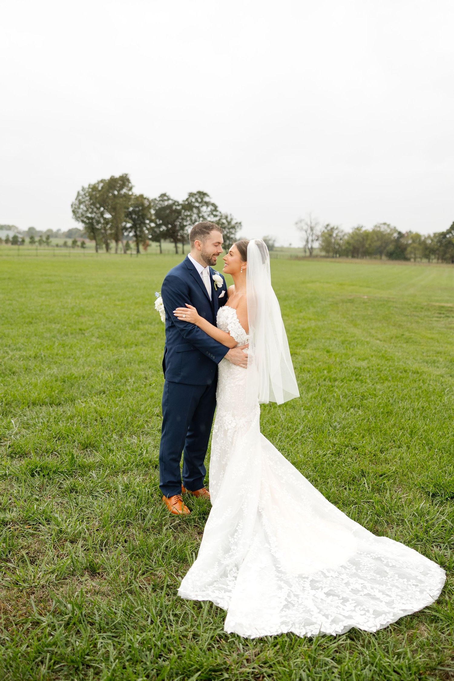 A romantic wedding couple embraces in a green field with a flowing white lace dress trailing behind them.