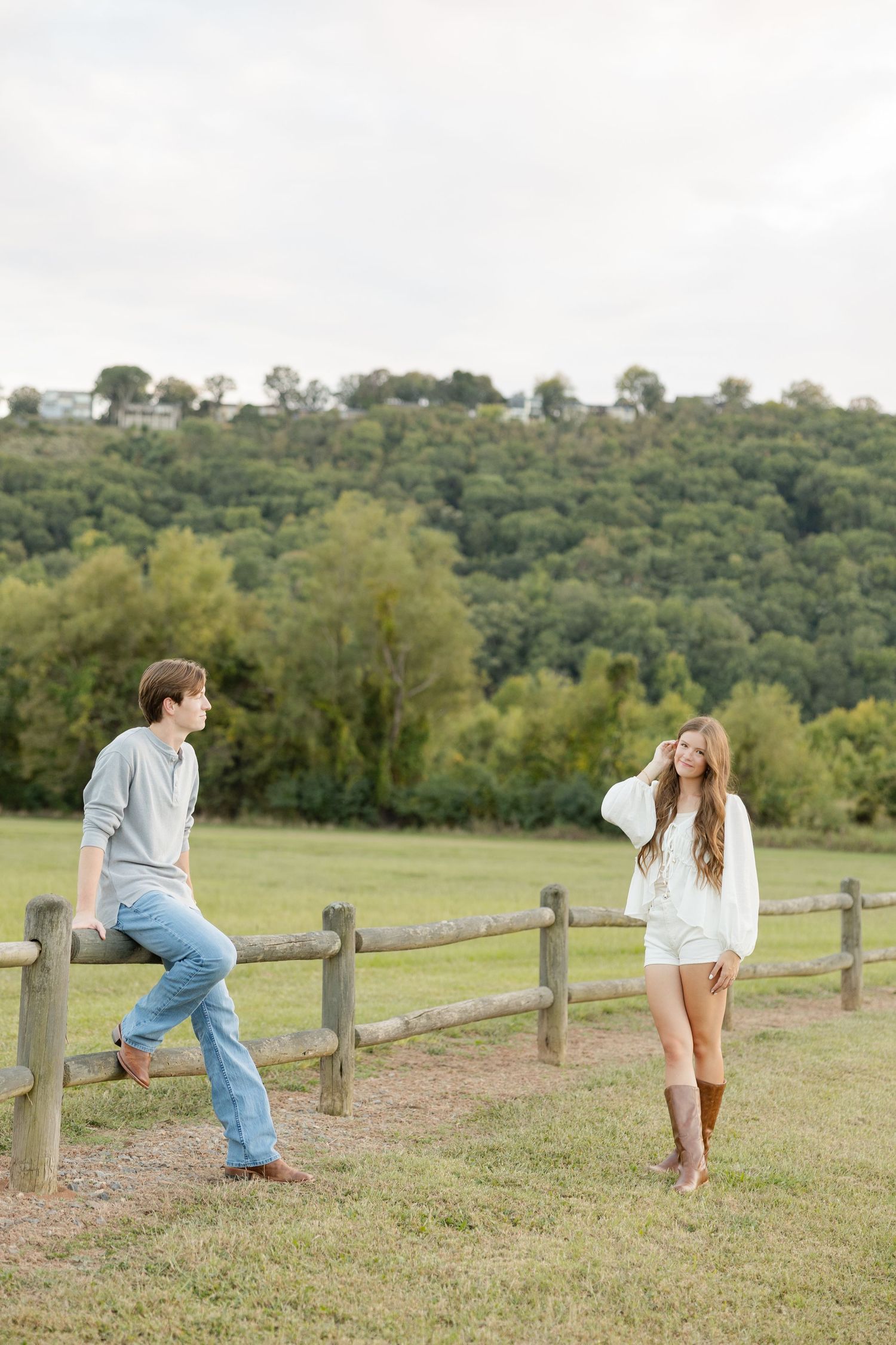 Playful couple interacting along a wooden fence in a scenic countryside meadow with rolling hills in the background.