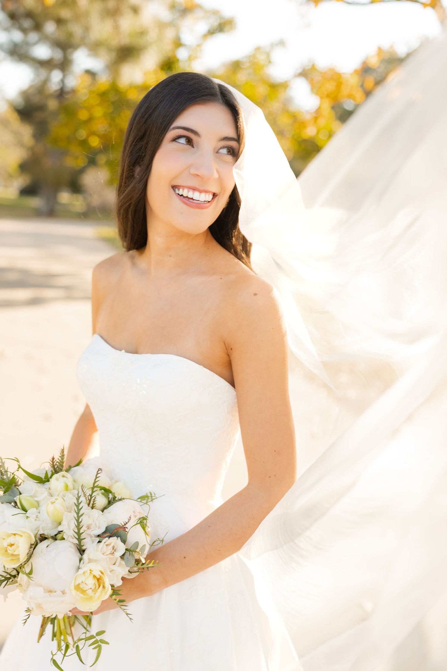 Bride with flowing veil smiles in sunlit outdoor portrait with bouquet.