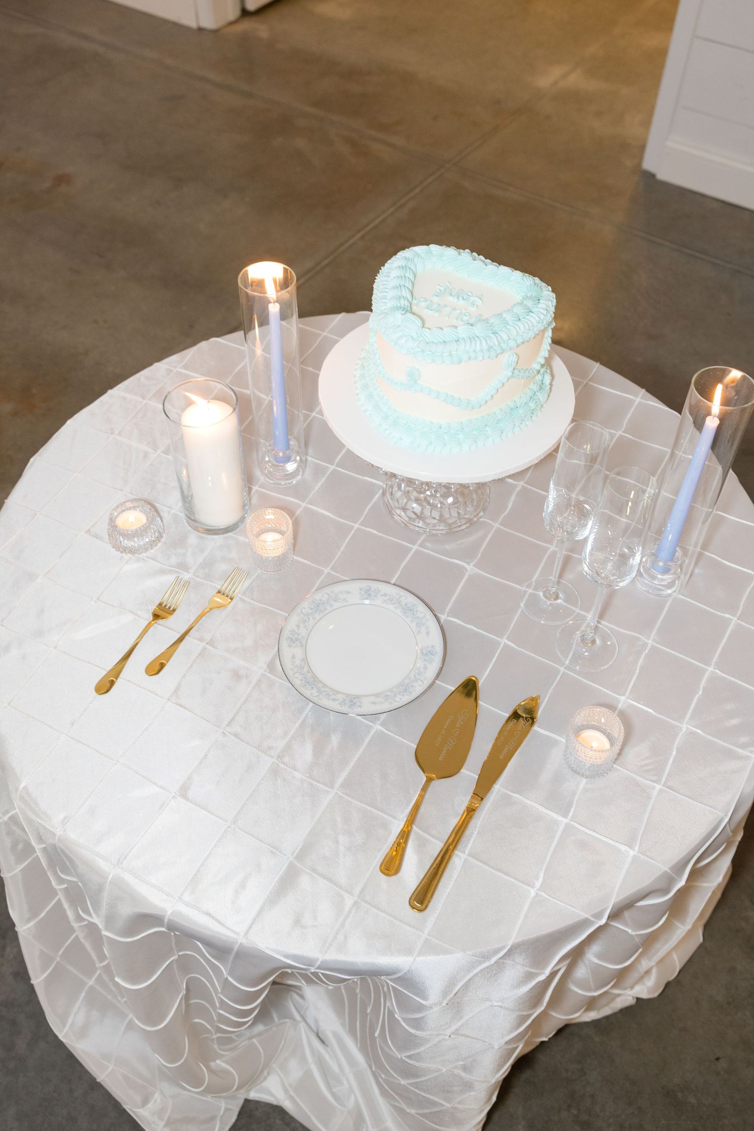 Elegant wedding cake table setting with light blue cake, gold utensils and white tablecloth.