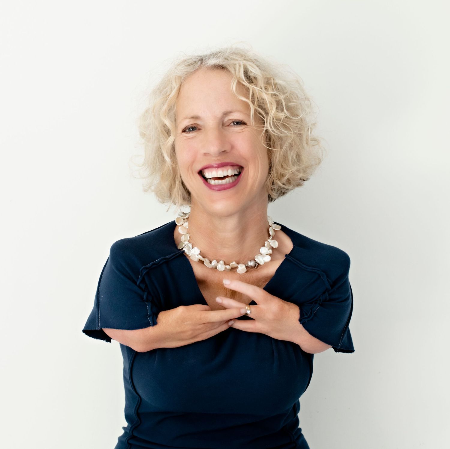 Laughing woman with curly blonde hair wears navy top and pearl necklace indoors.