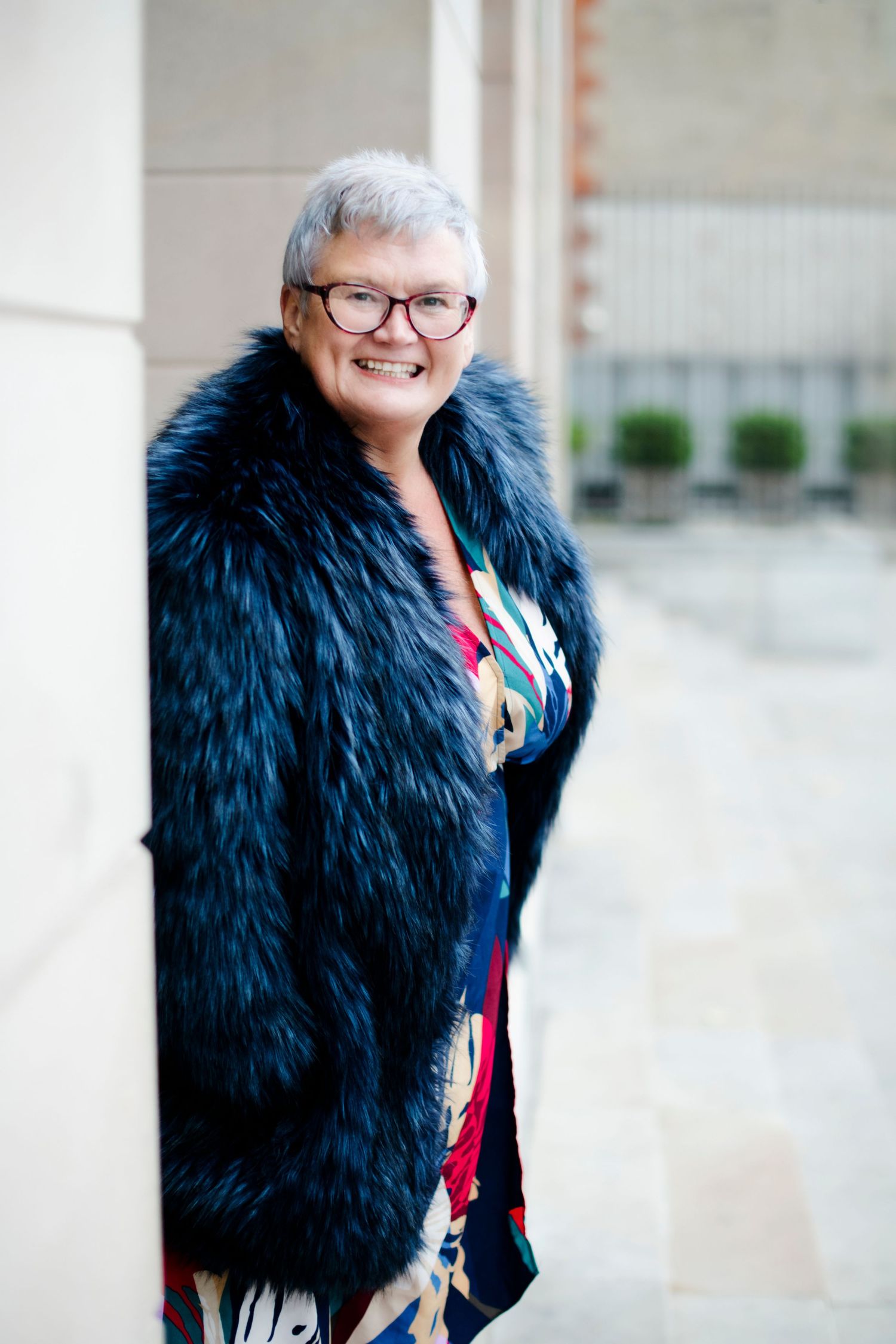 Smiling older woman with gray hair and glasses wearing a black fur coat leaning against a stone pillar.