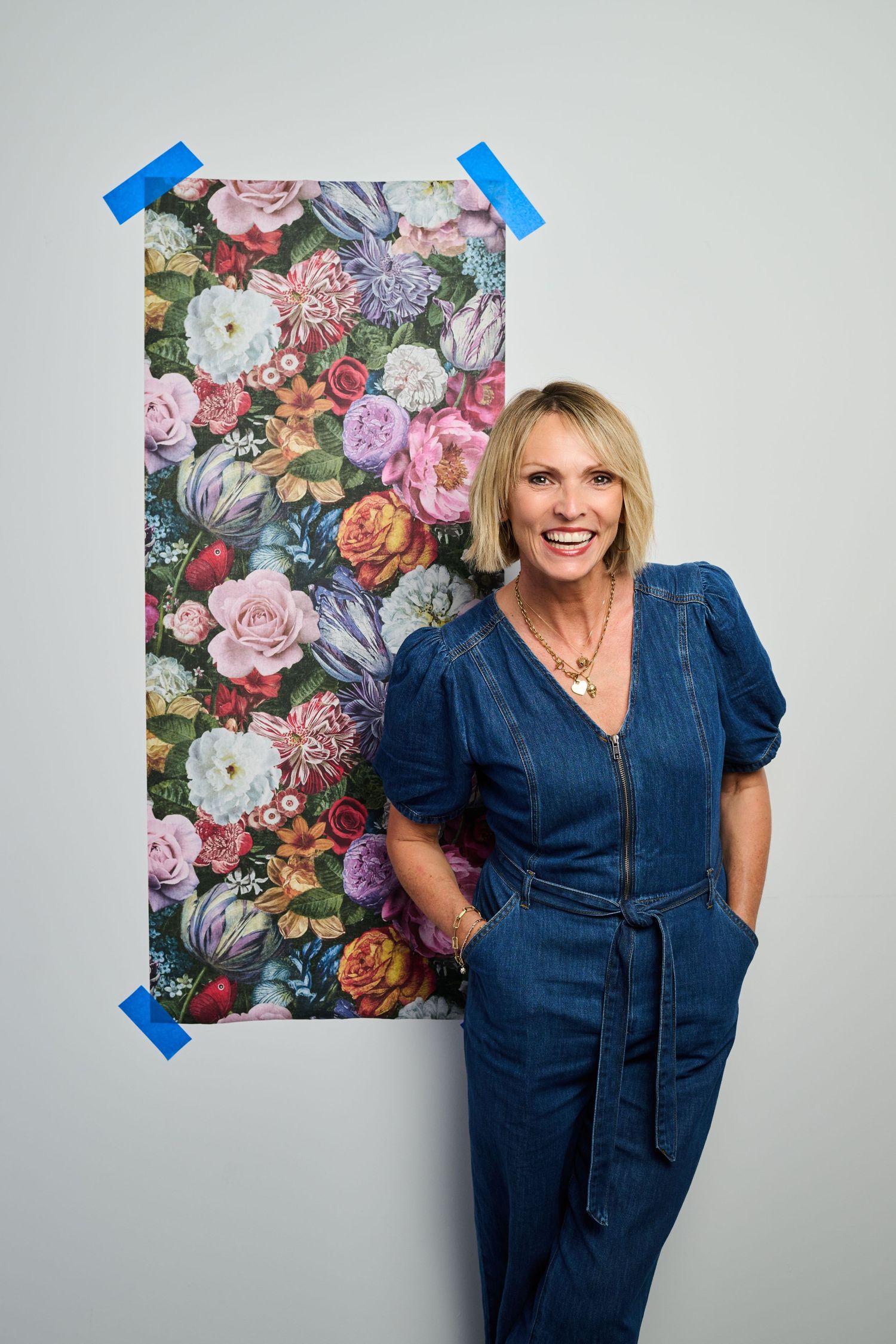 Happy blonde woman in blue denim jumpsuit standing beside vibrant floral wallpaper sample taped to white wall.
