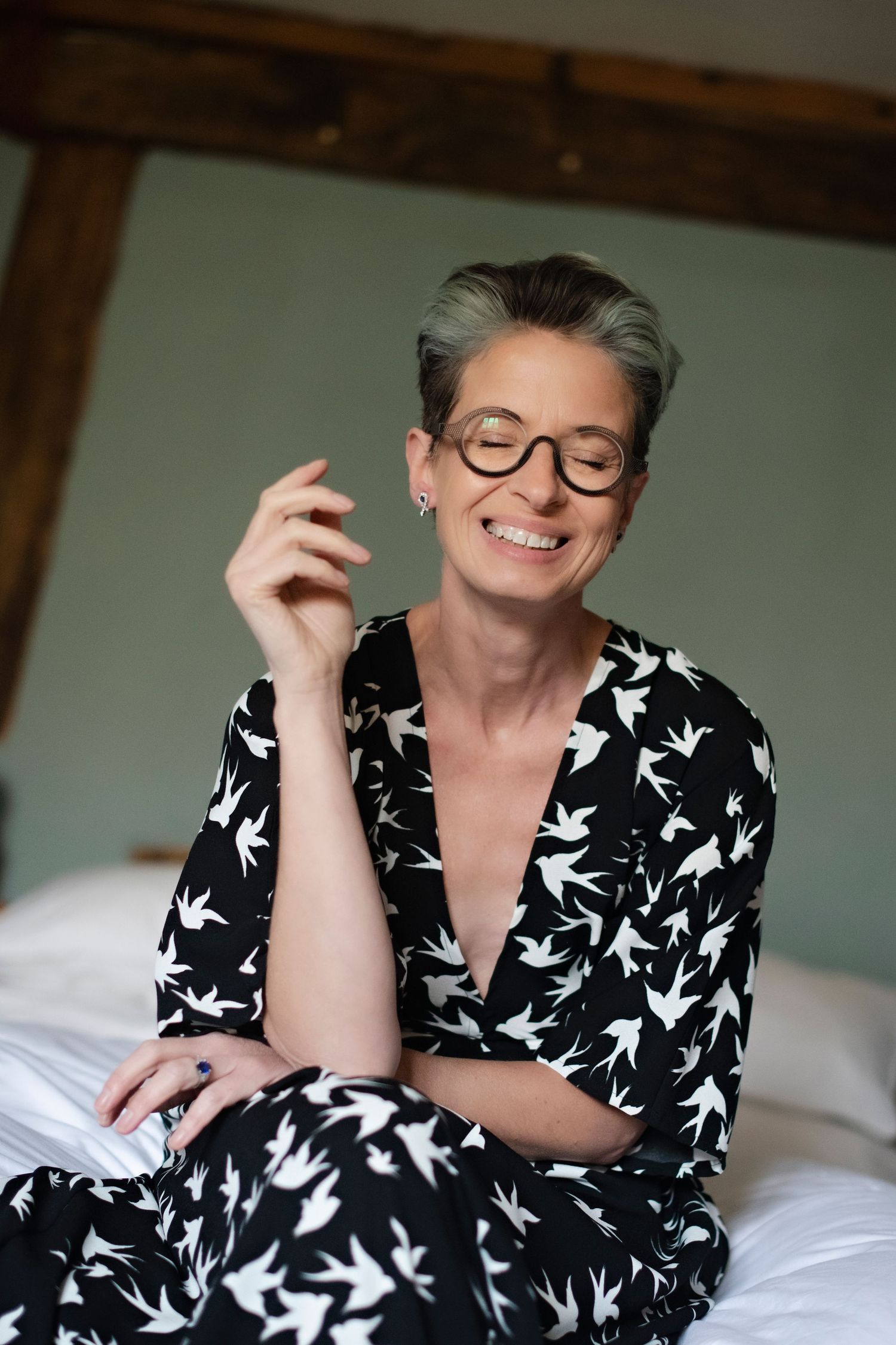 Smiling woman with short gray hair and round glasses in a black and white bird-print dress sitting on a bed.