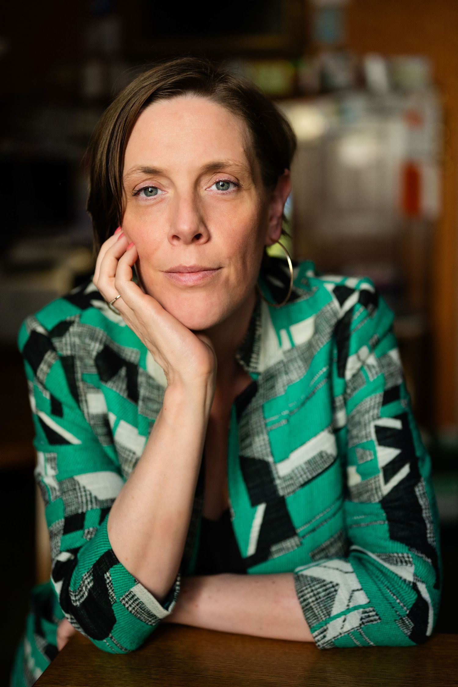 Woman in green geometric blazer rests head on hand at a table in a warmly lit interior.