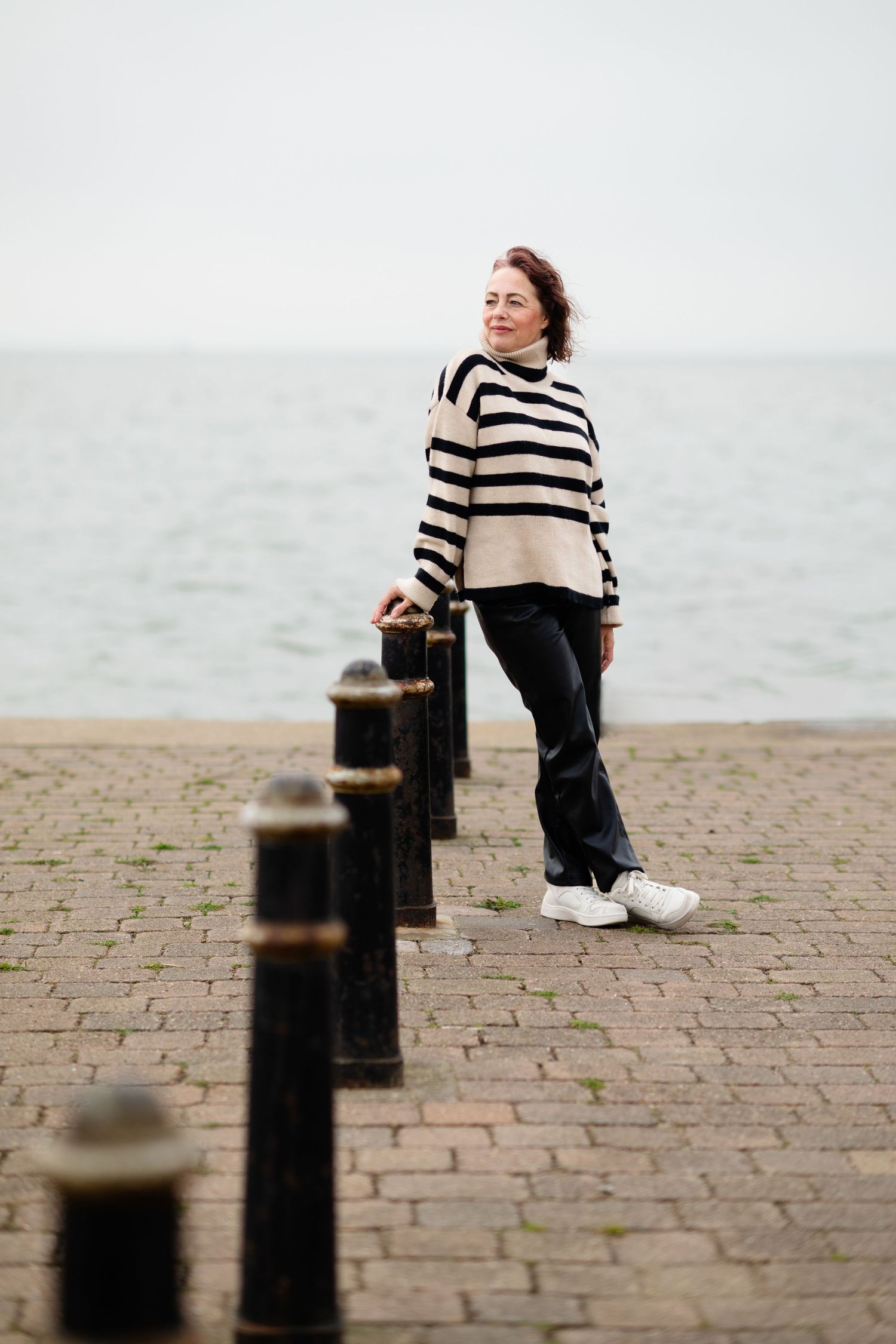 Woman in striped turtleneck sweater standing by bollards on a waterfront promenade.