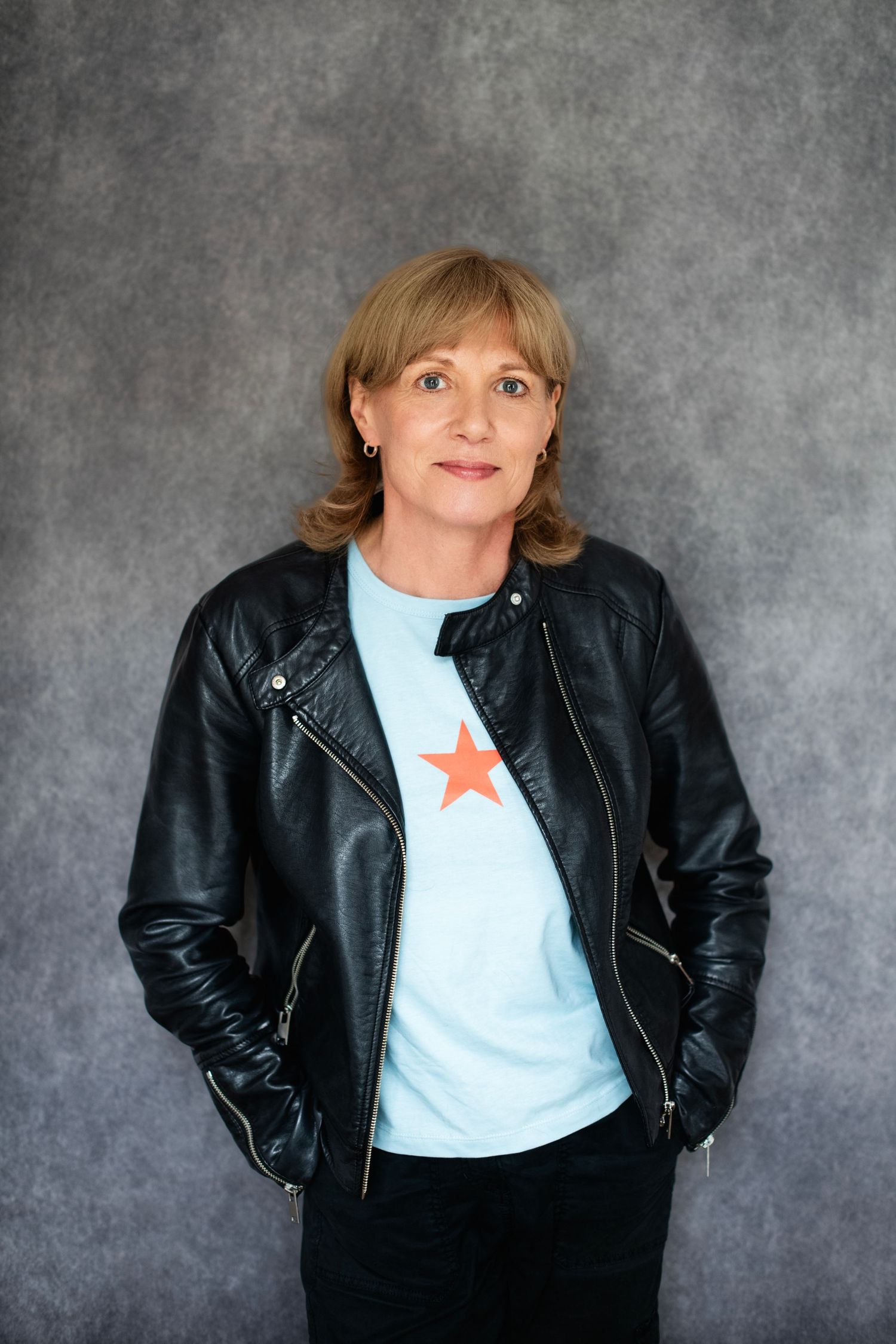 Woman in black leather jacket and light blue star t-shirt standing with hands in pockets against gray background.