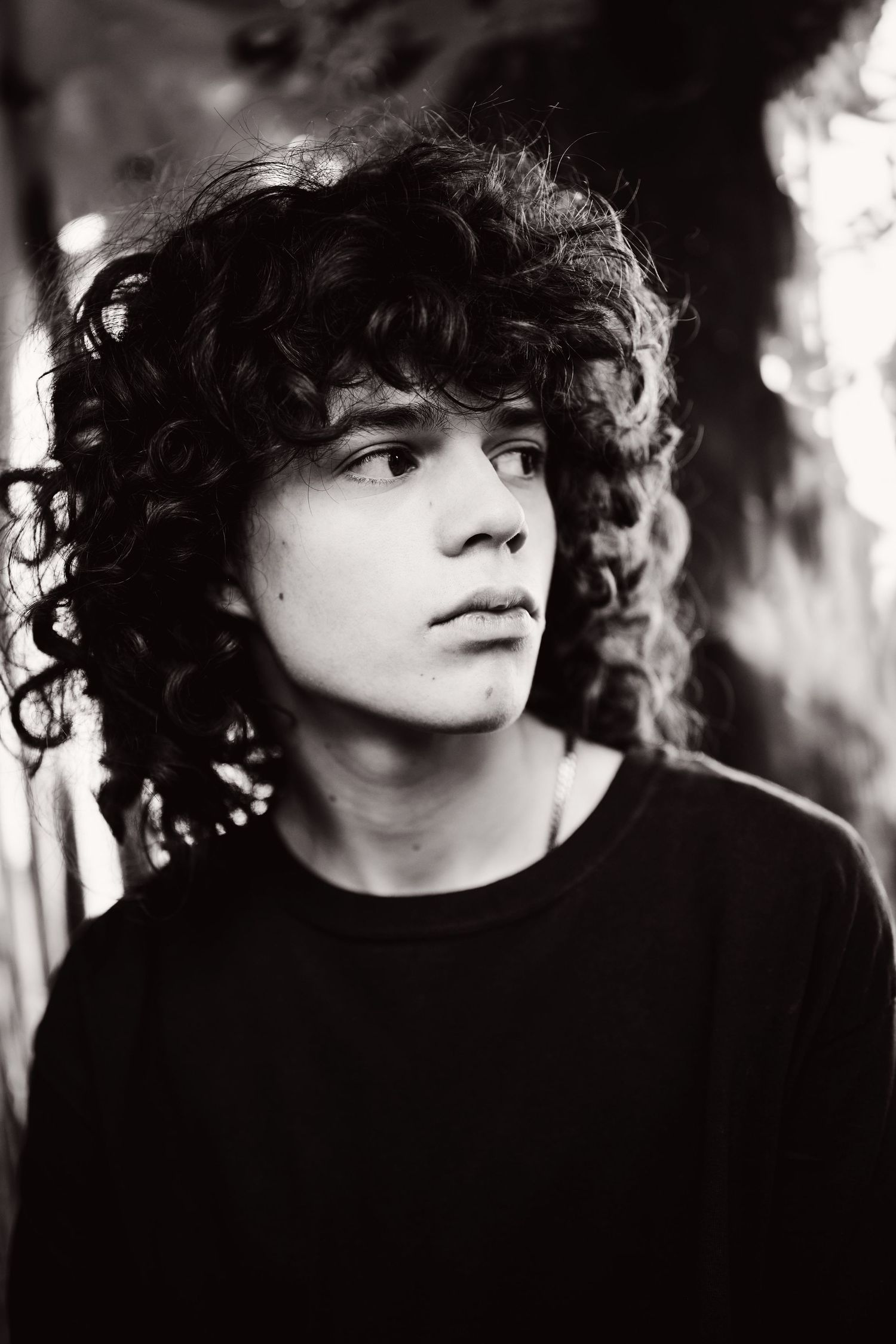 Black and white portrait of a young man with long curly hair wearing a black shirt, looking thoughtfully to the side.