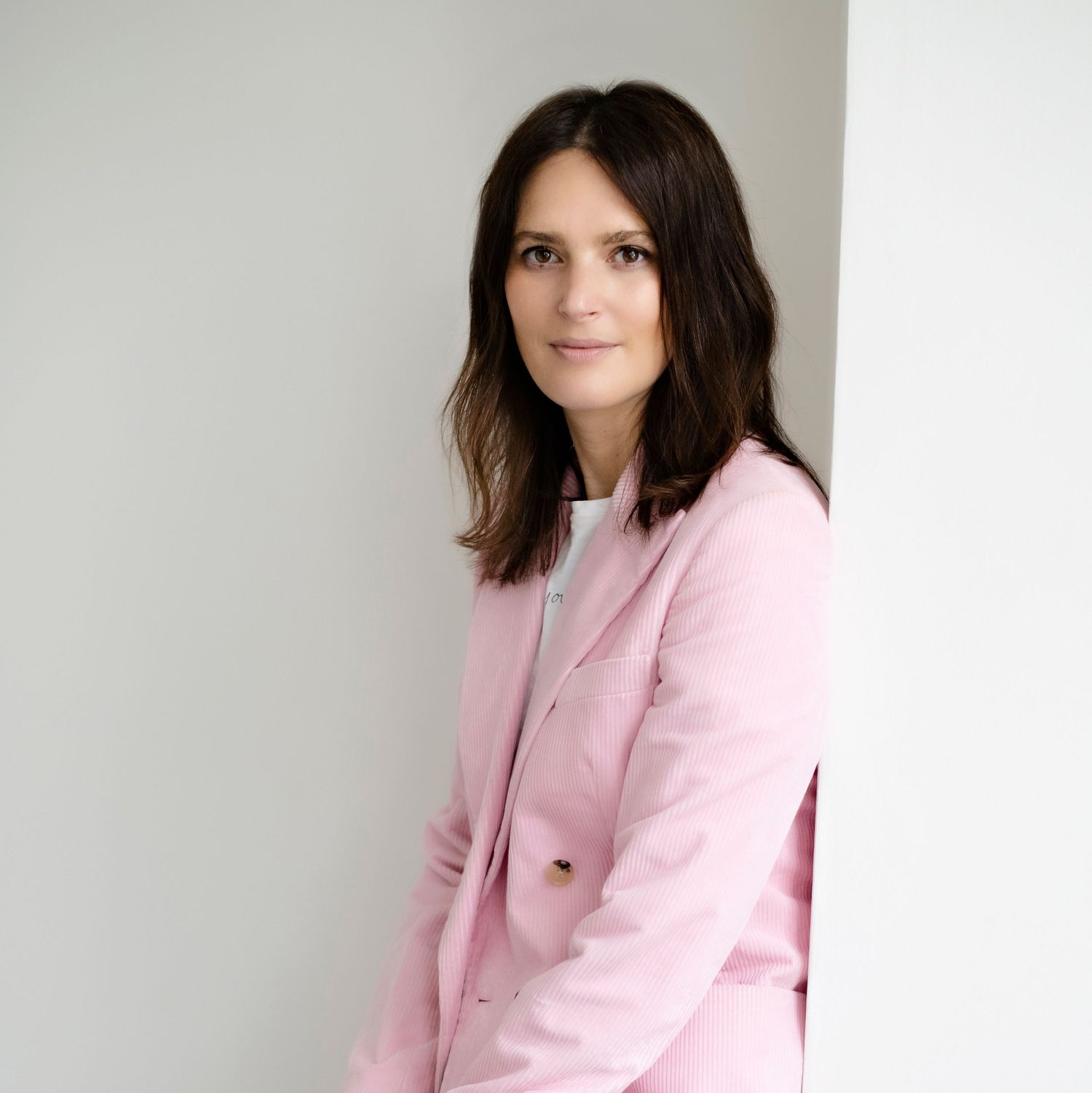 Dark-haired woman in a pink blazer leaning against a white wall in a professional portrait.