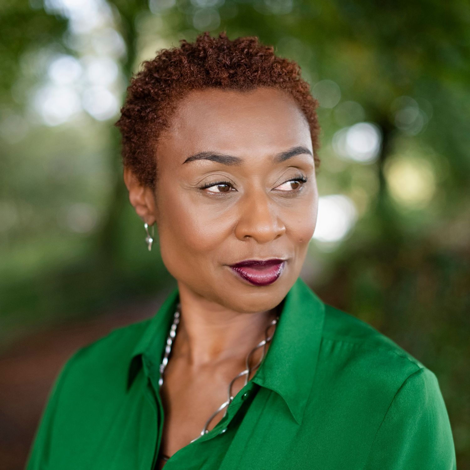 Black woman with short natural auburn hair in a green shirt posing outdoors among green trees.