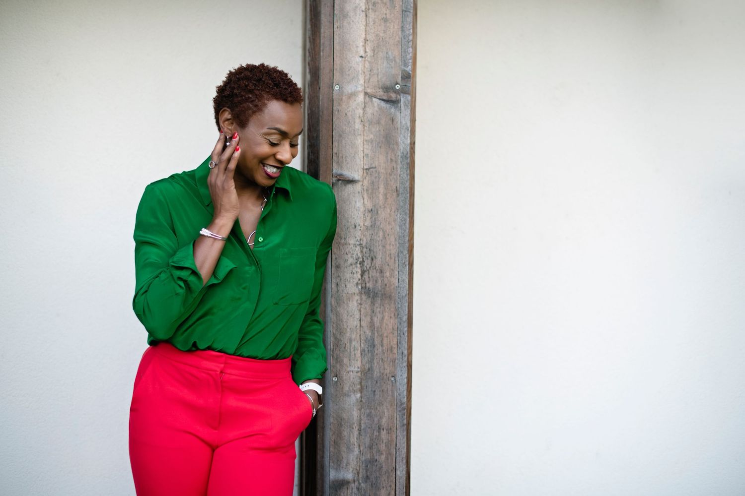 Black woman in green shirt and red pants leaning against a wooden post smiling downward in natural light.