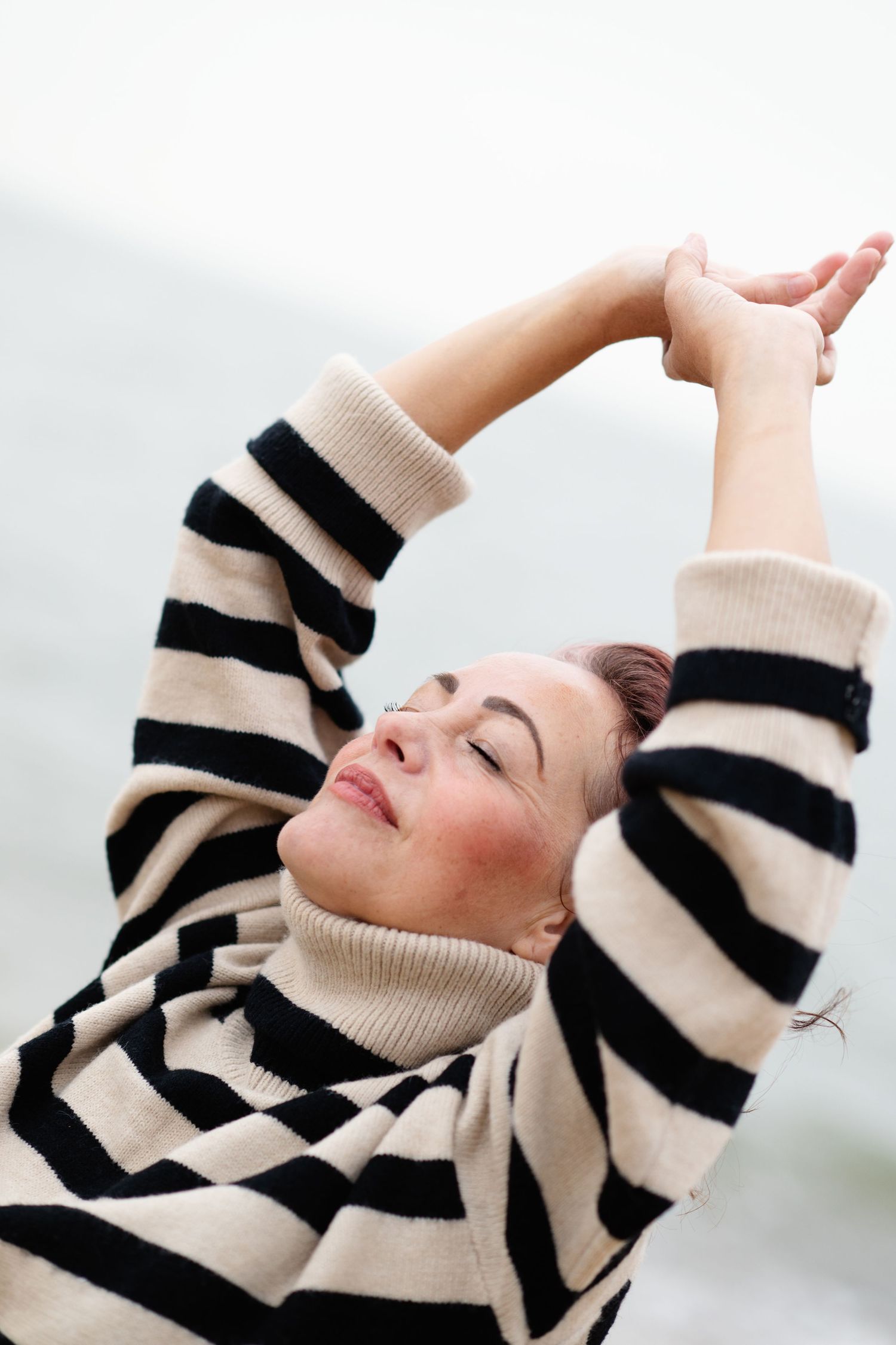 A woman in a striped turtleneck sweater stretches her arms overhead with eyes closed outdoors.