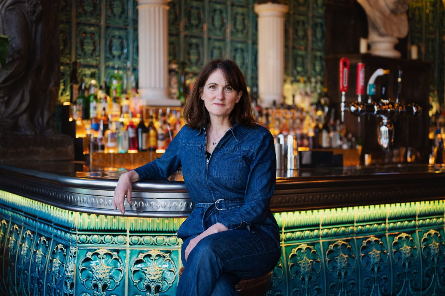 Woman in denim jumpsuit sitting at elegant green tiled bar with backlit bottle display.