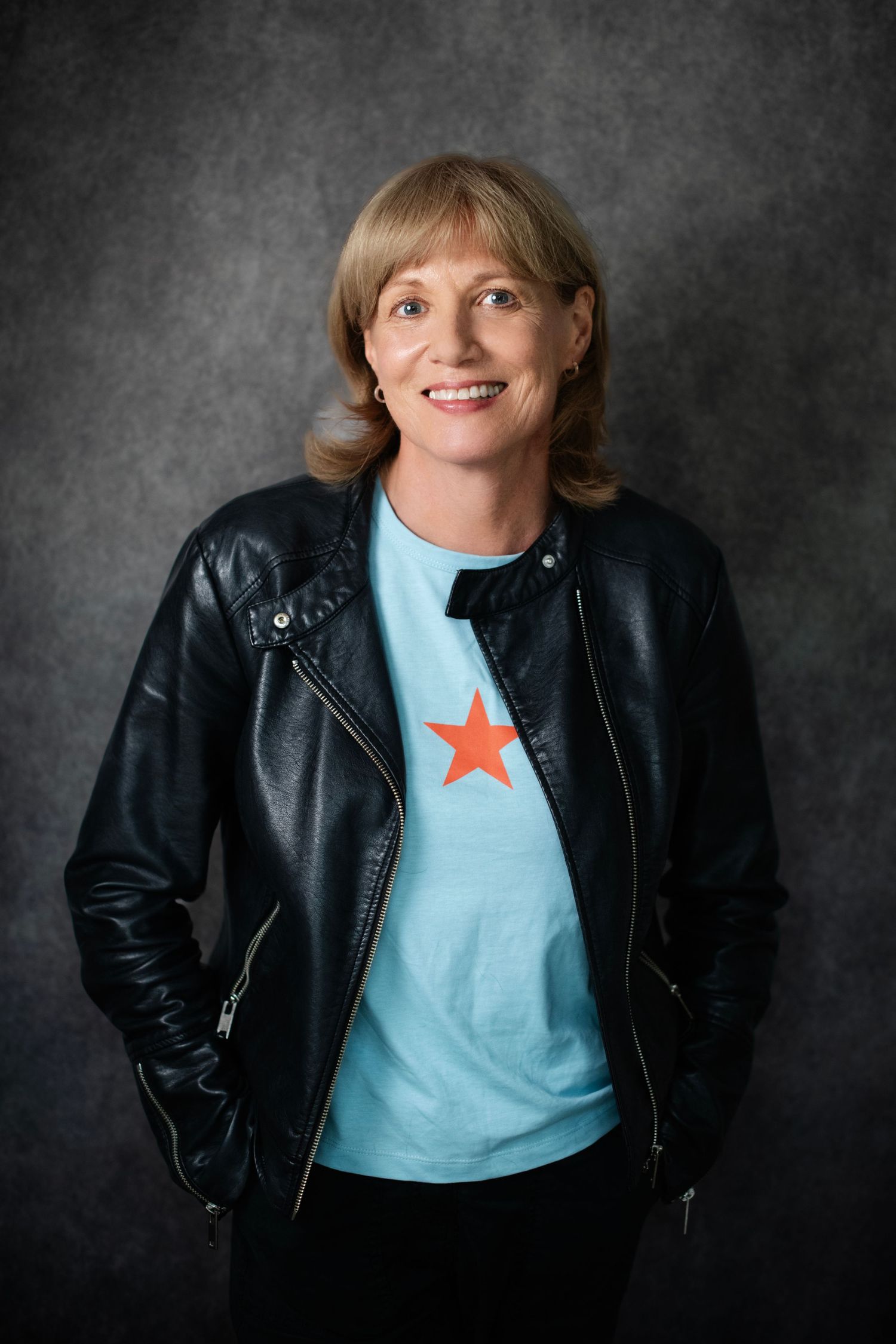 Smiling woman in black leather jacket and light blue star print t-shirt posing confidently against gray backdrop.