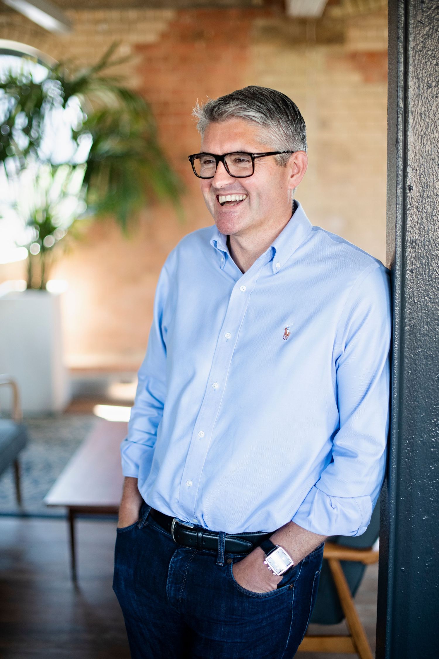 Laughing man in light blue shirt leaning against wall in bright office space.