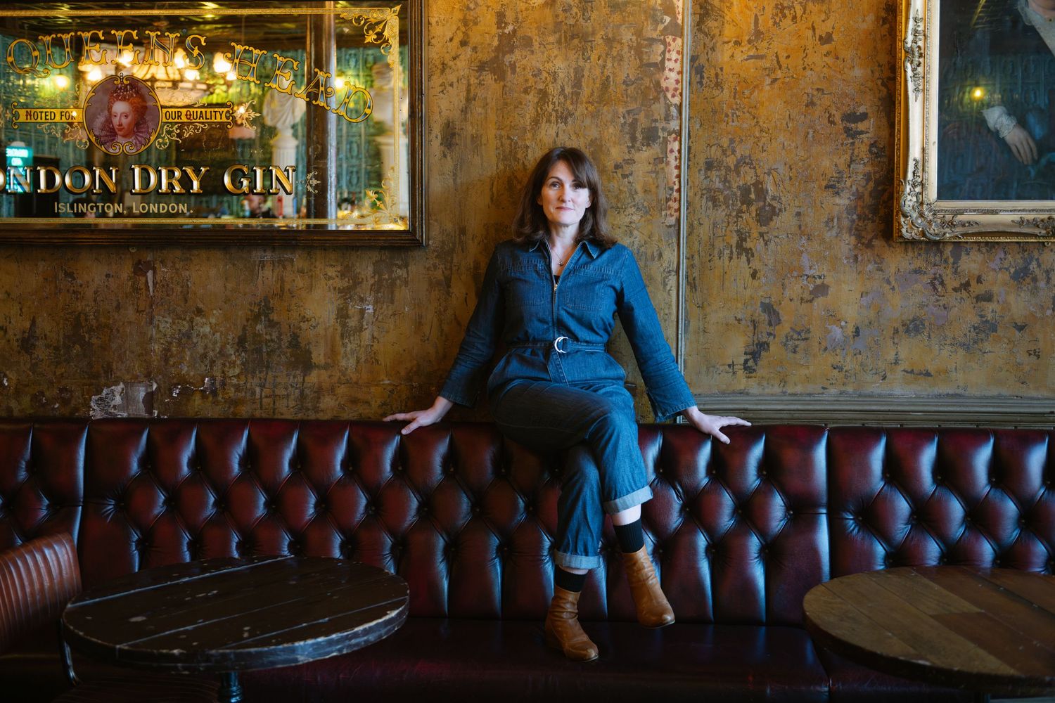 A stylish woman in a denim jumpsuit sits on a leather Chesterfield sofa in a vintage bar setting.