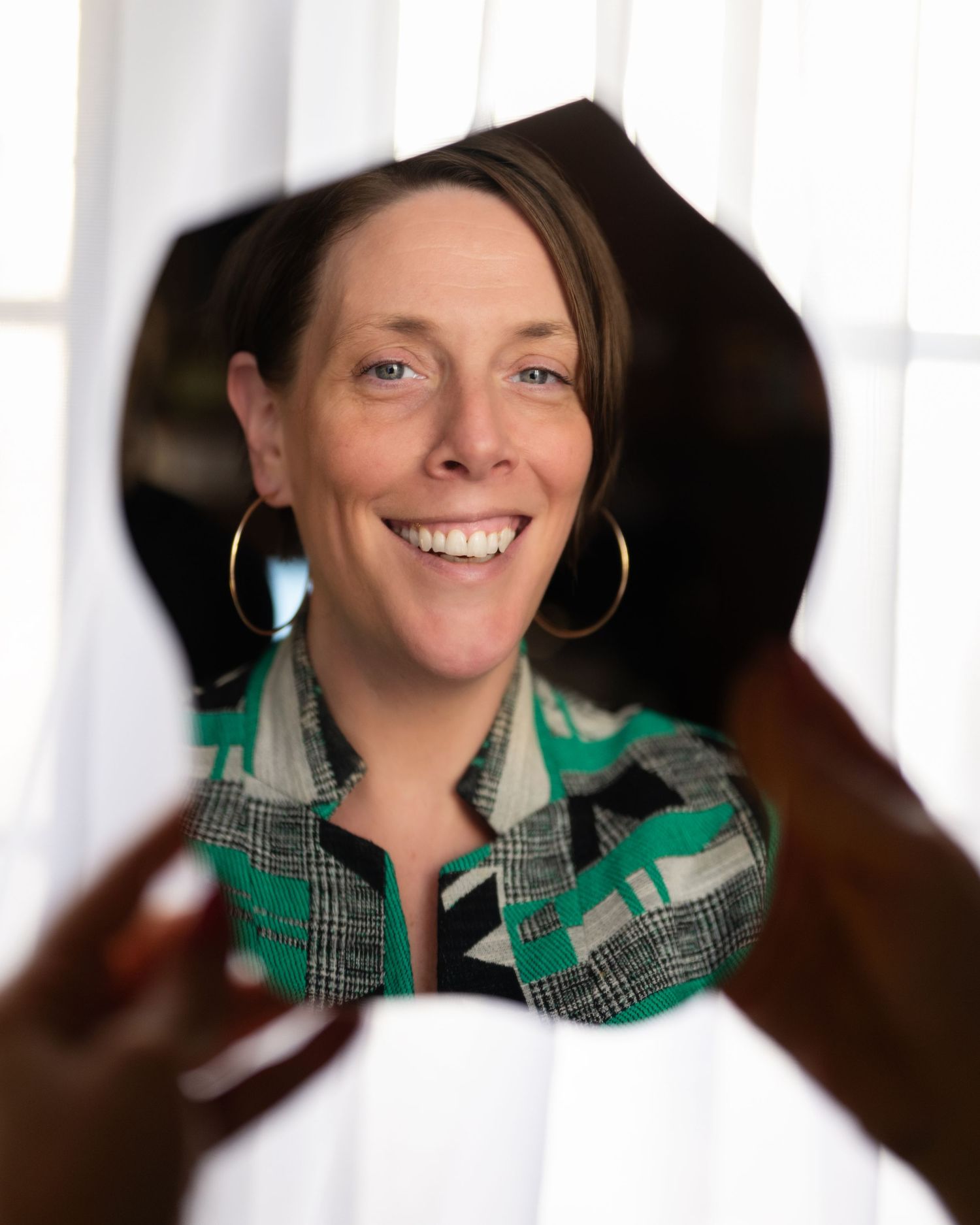 Smiling woman in green patterned jacket wearing gold hoop earrings reflected in a handheld mirror.
