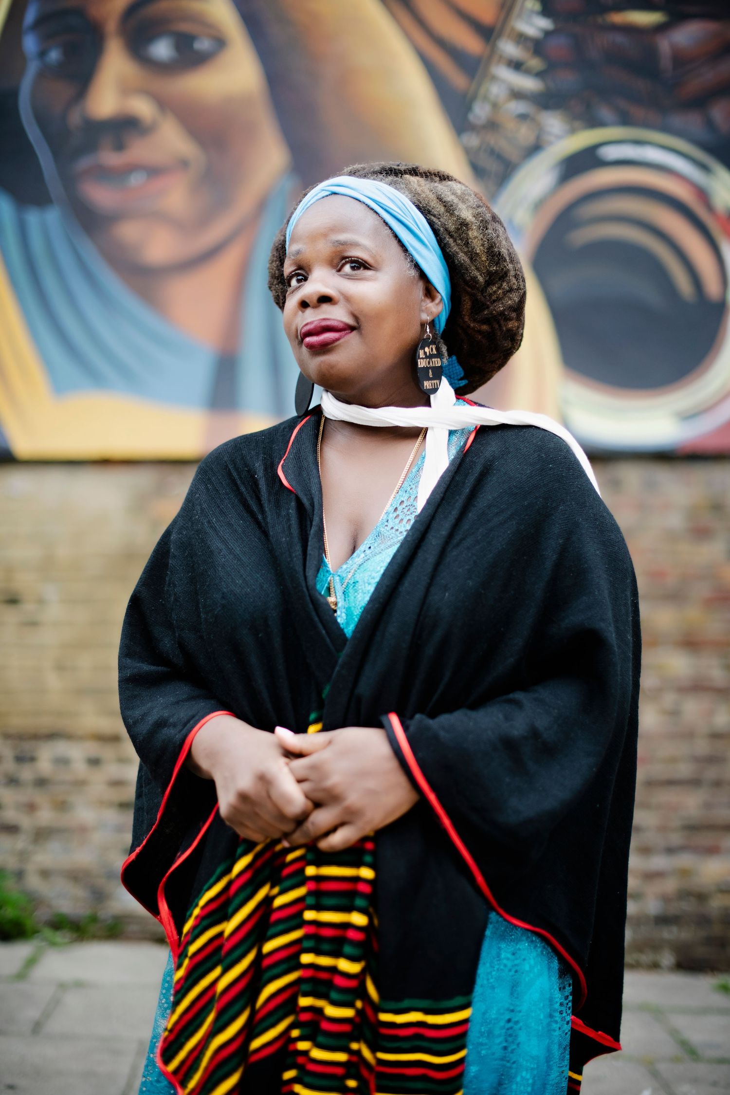 Woman in black cape with kente cloth accents stands before a vibrant mural wearing a blue headband and matching dress.