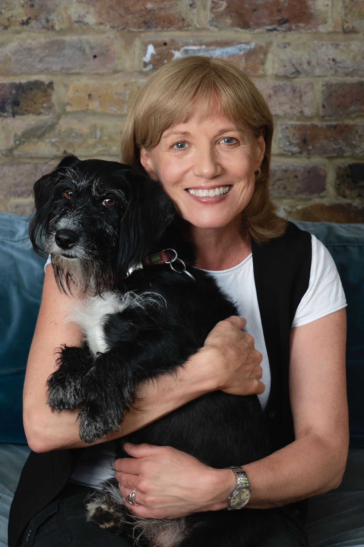 Smiling woman holding a fluffy black and white dog while seated on teal sofa against brick wall.