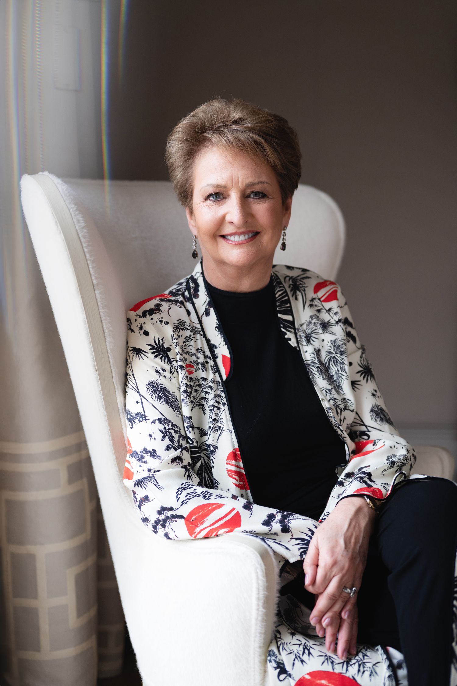 Smiling woman in floral jacket seated comfortably in white armchair near draped window.