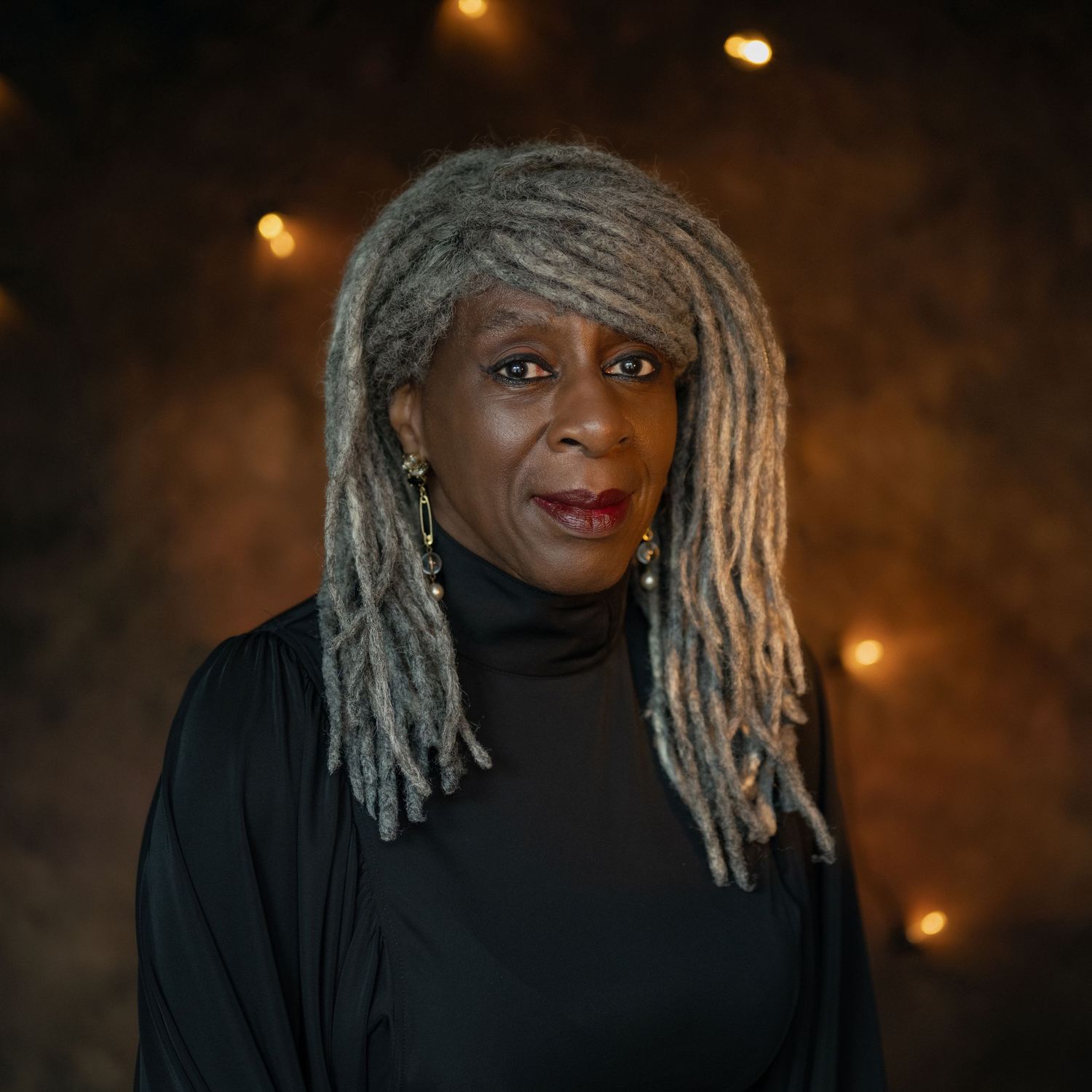 A Black woman with long gray locs wearing a black turtleneck poses against a dark bokeh background.