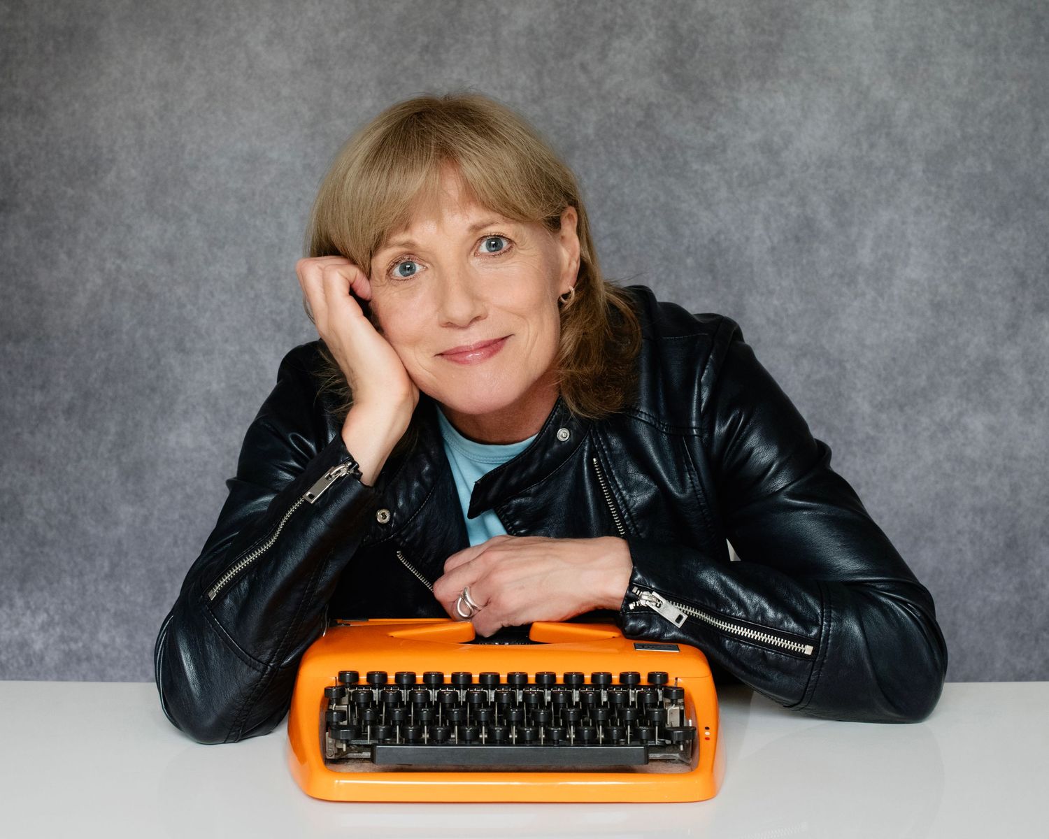 Woman in leather jacket resting head on hand beside vintage orange typewriter on white table.