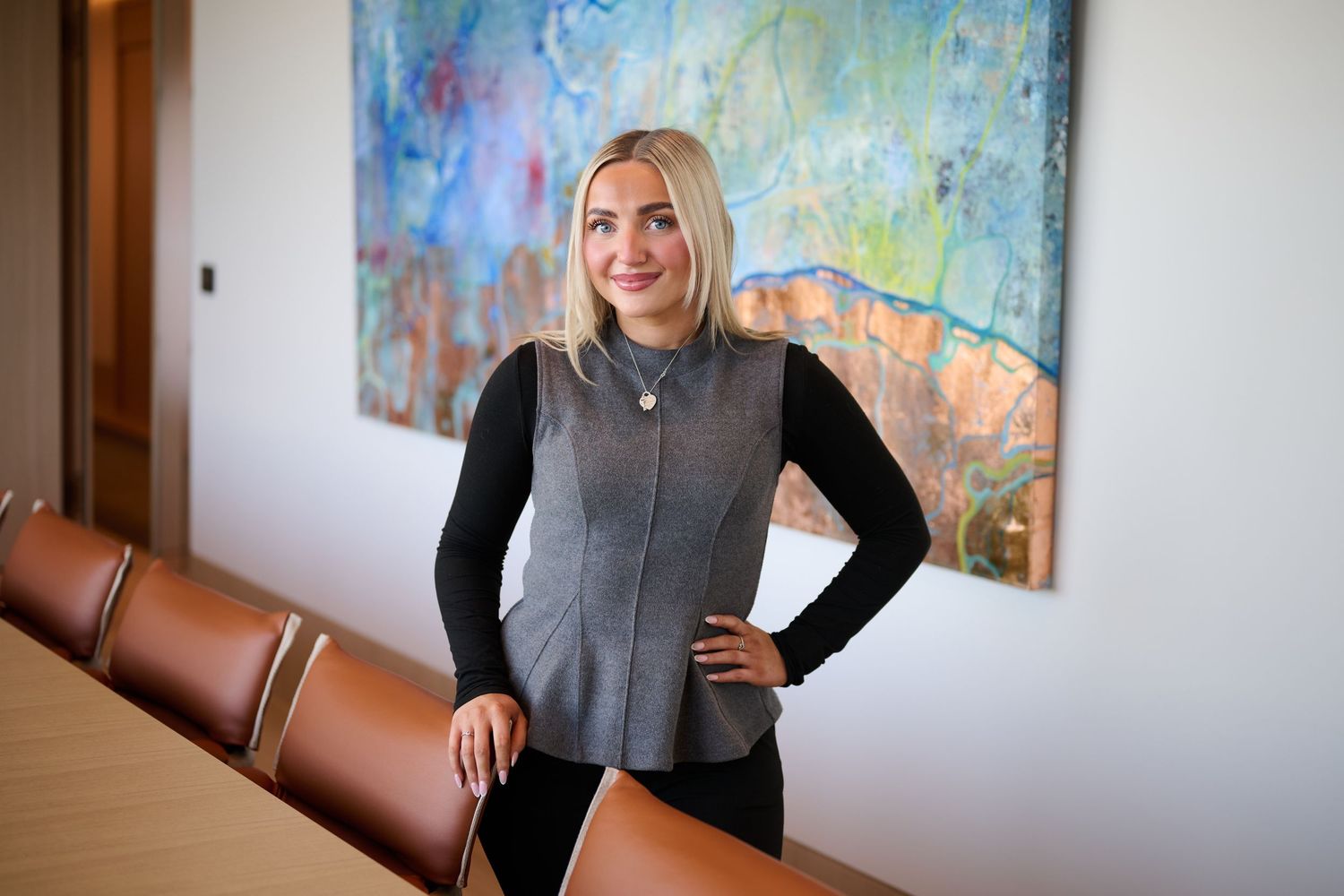 Professional woman in gray vest poses confidently in modern boardroom with colorful abstract artwork.