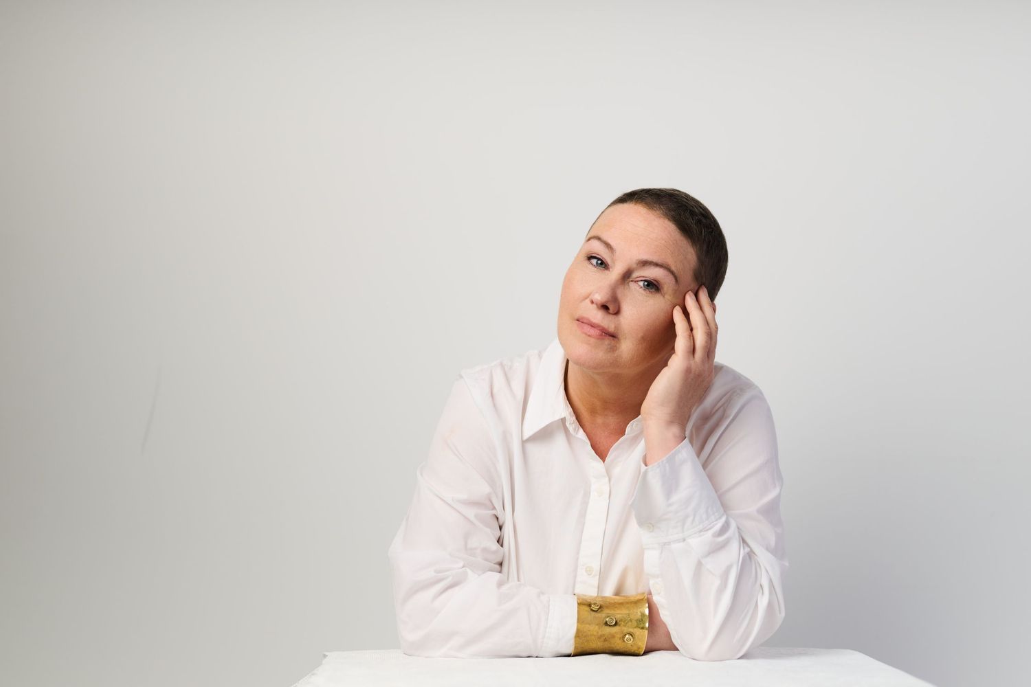 Woman in white shirt sitting at table, resting head on hand, looking up thoughtfully against white background.