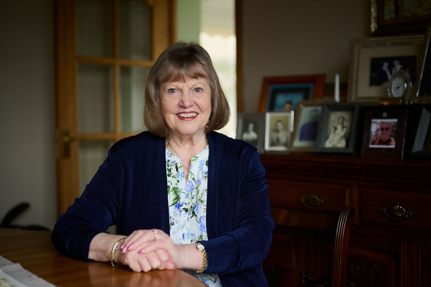 Smiling senior woman in blue blazer sits at wooden desk in a warmly decorated home office.