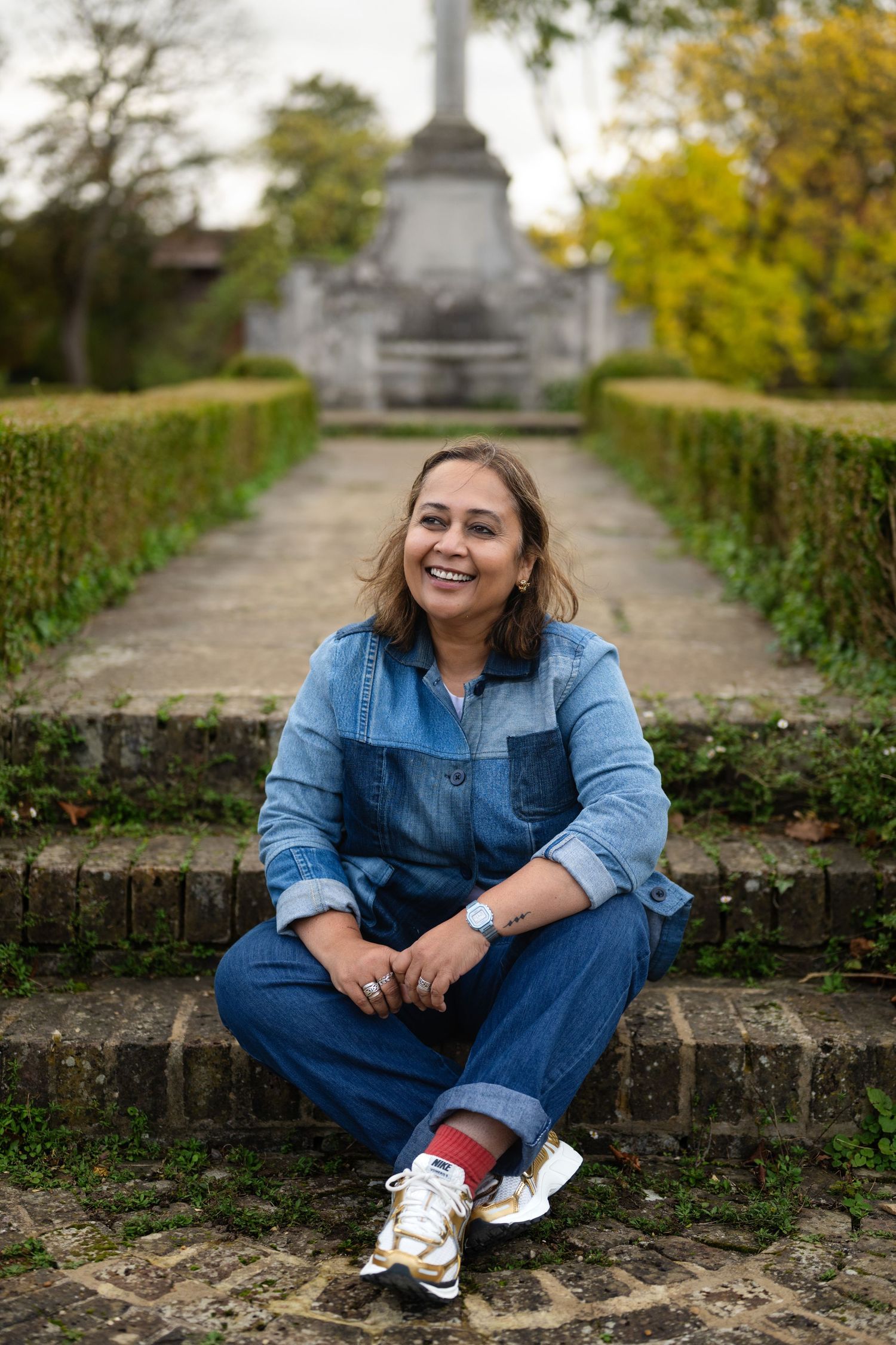 Smiling woman in denim jumpsuit sitting on stone steps in an autumn garden with a monument.