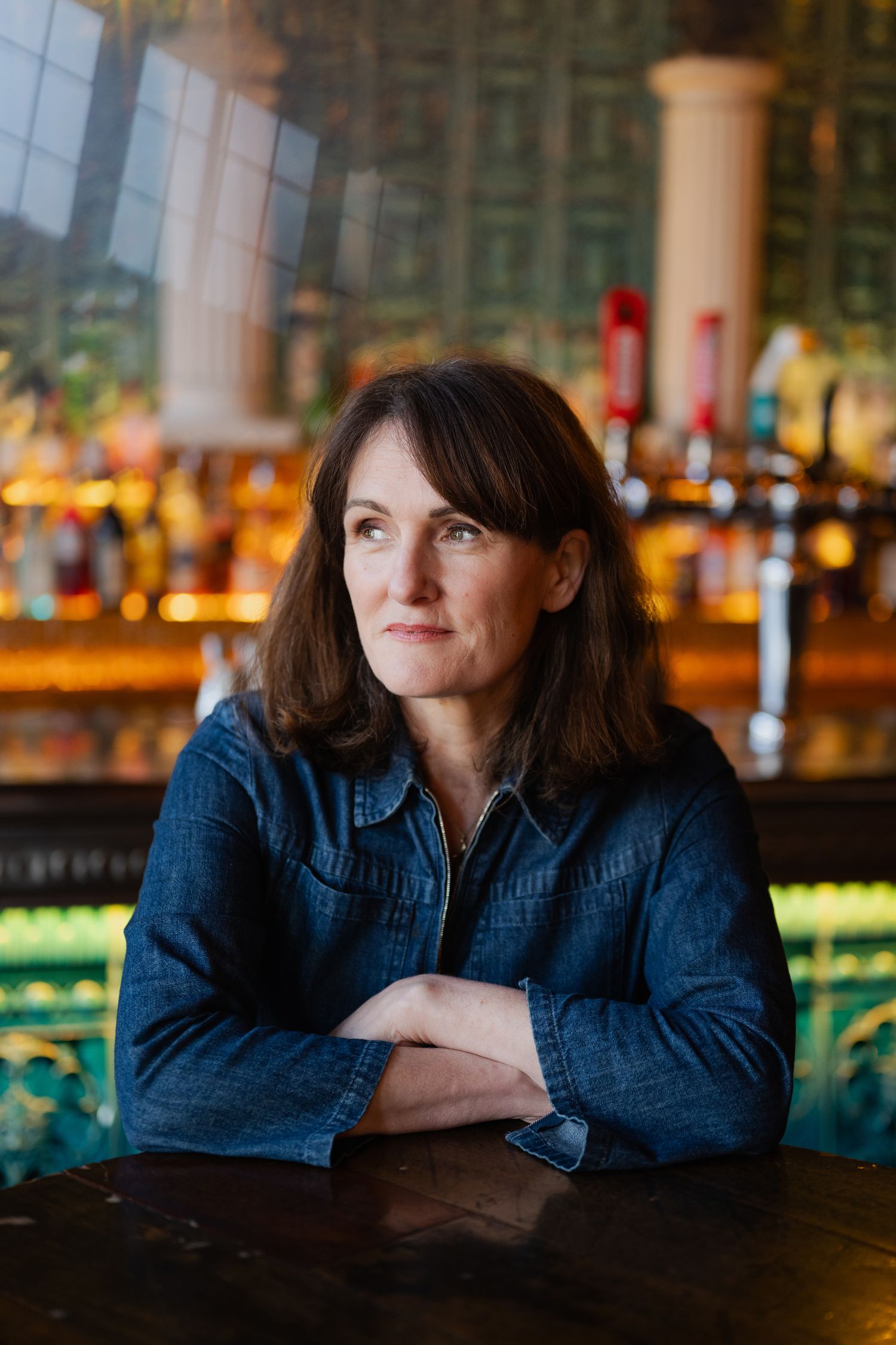 Woman in denim jacket sitting at bar with arms crossed, looking thoughtfully to the side.
