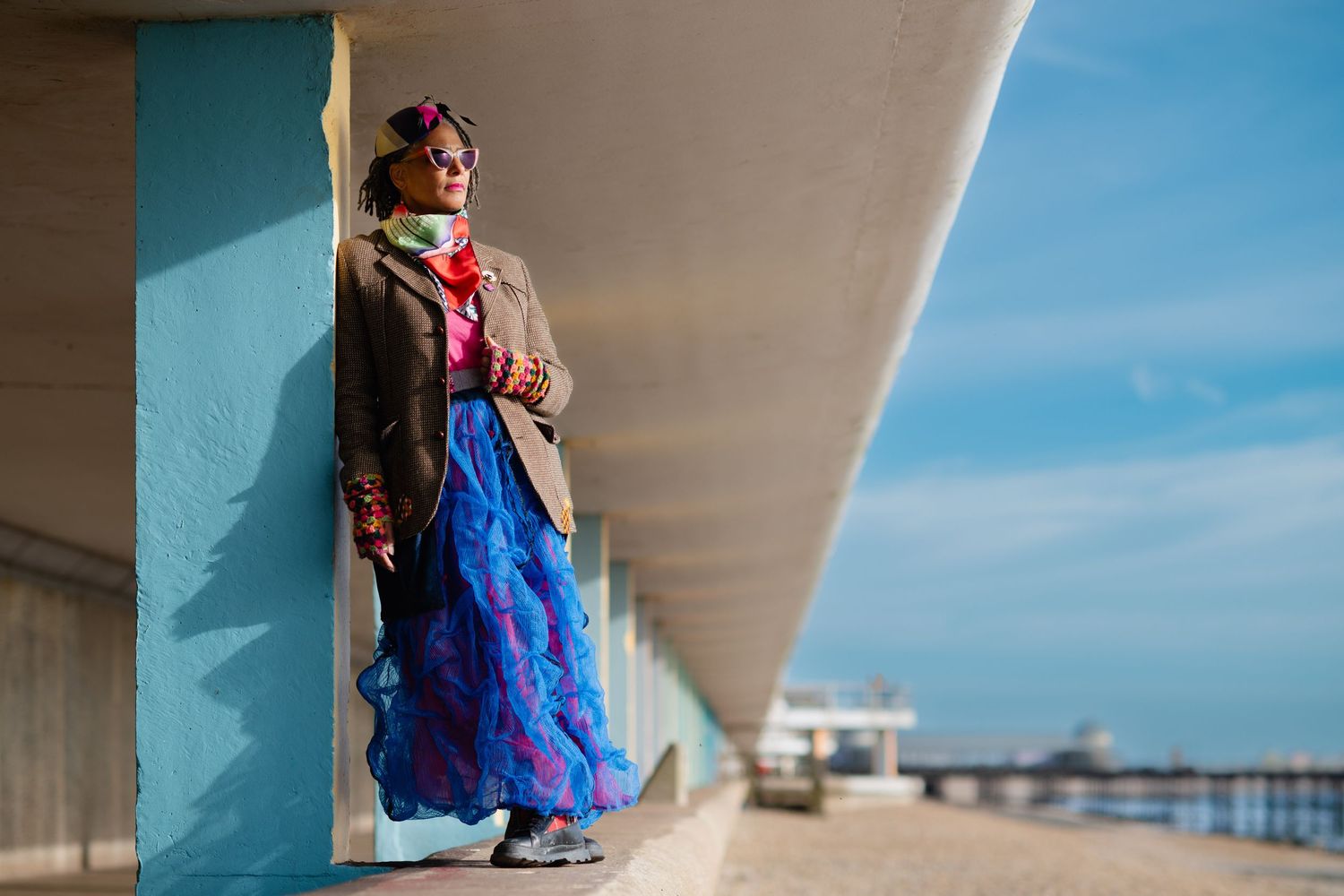 A woman in a colorful blue skirt and brown jacket leans against a pastel wall on a sunny seaside promenade.