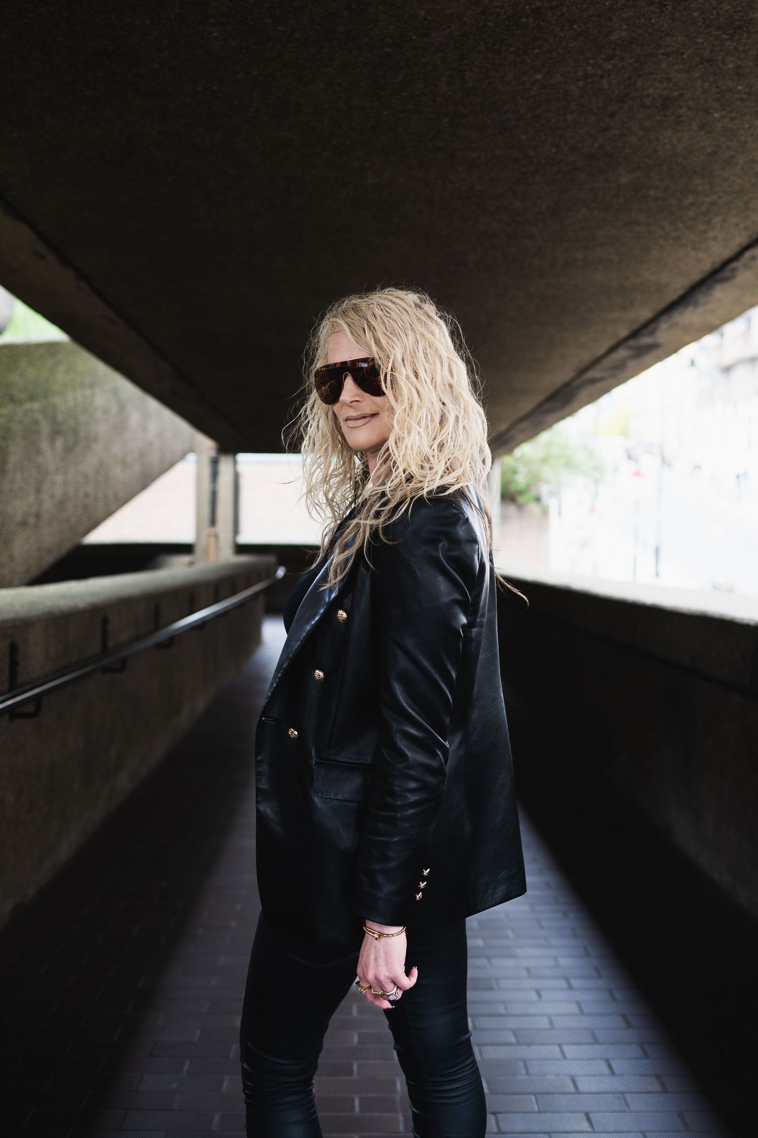 Blonde woman in black leather jacket and sunglasses walking through an urban underpass corridor.