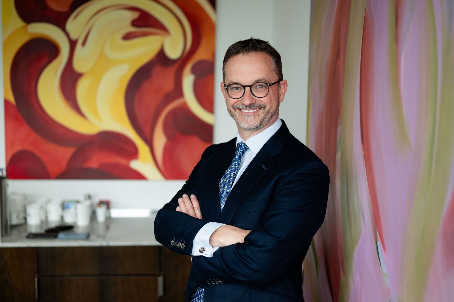 Smiling businessman in navy suit with arms crossed standing near colorful artwork.