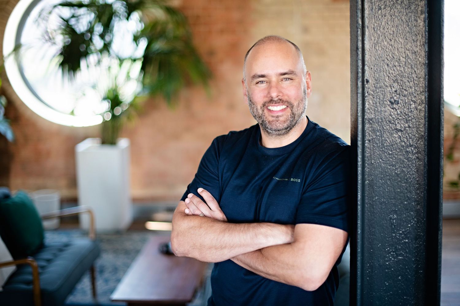 Bearded man with crossed arms smiling in contemporary brick office space.