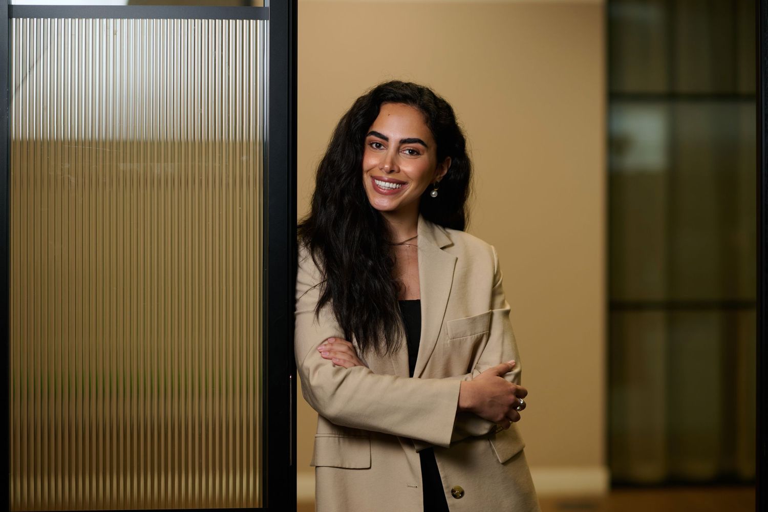 Professional woman in beige blazer smiling confidently, leaning against a glass office partition.