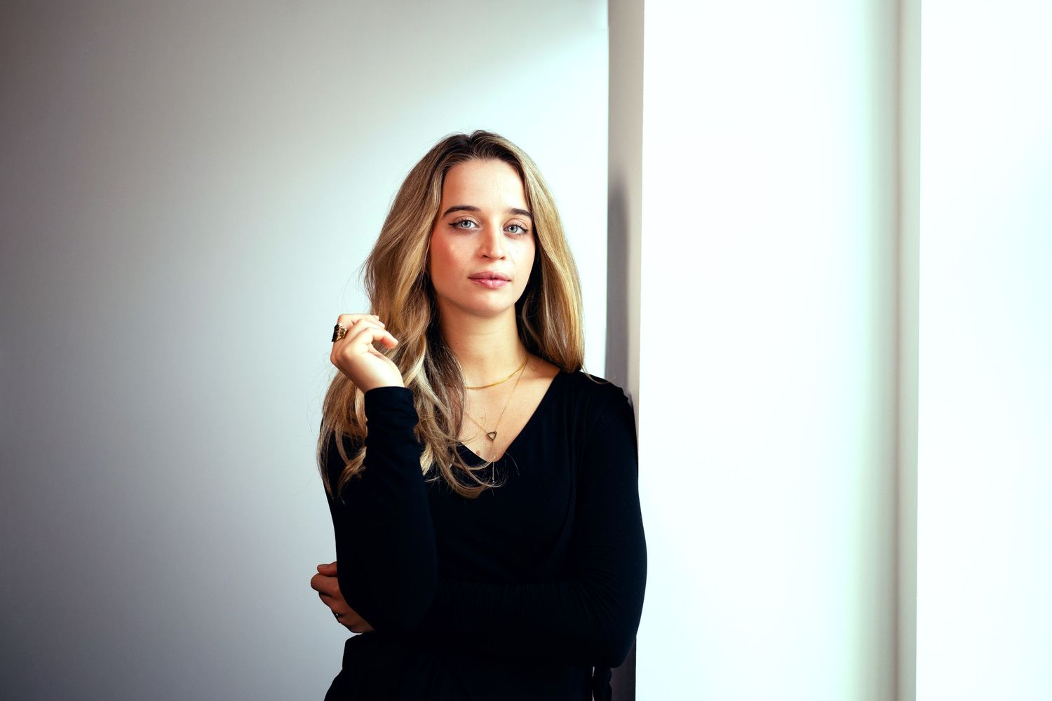 Young woman with long blonde hair in black top, arms crossed, posing thoughtfully near a bright window.