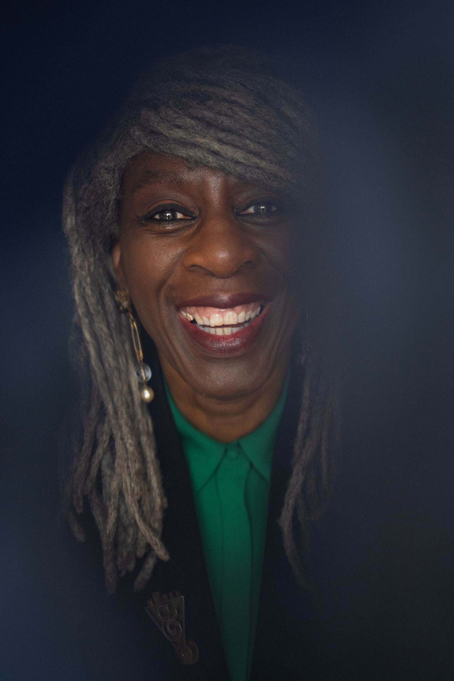 A joyful Black woman with gray locs laughs brightly while wearing a green shirt and gold brooch.