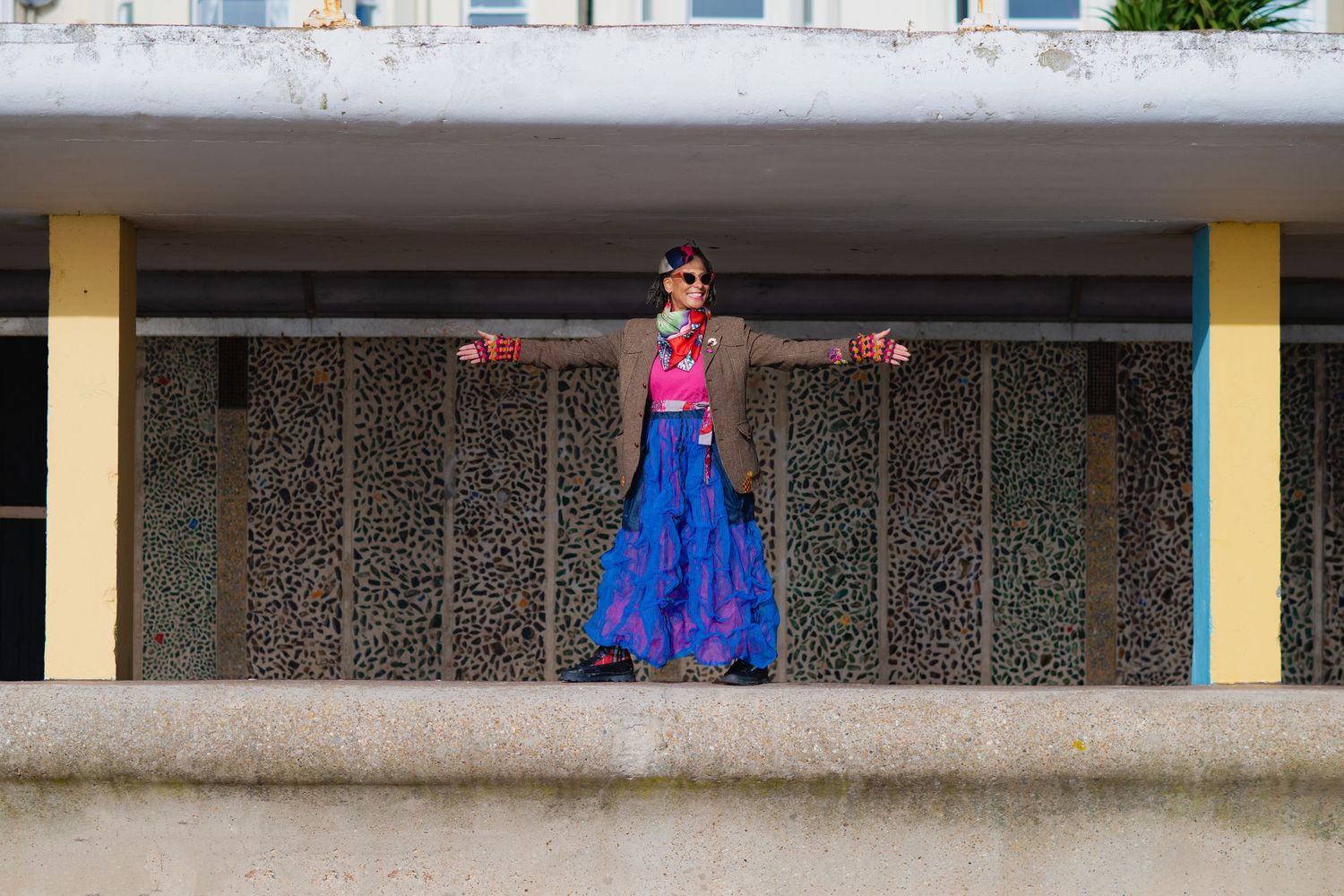 A woman in a vivid blue skirt and brown jacket stands with arms outstretched on an urban structure.