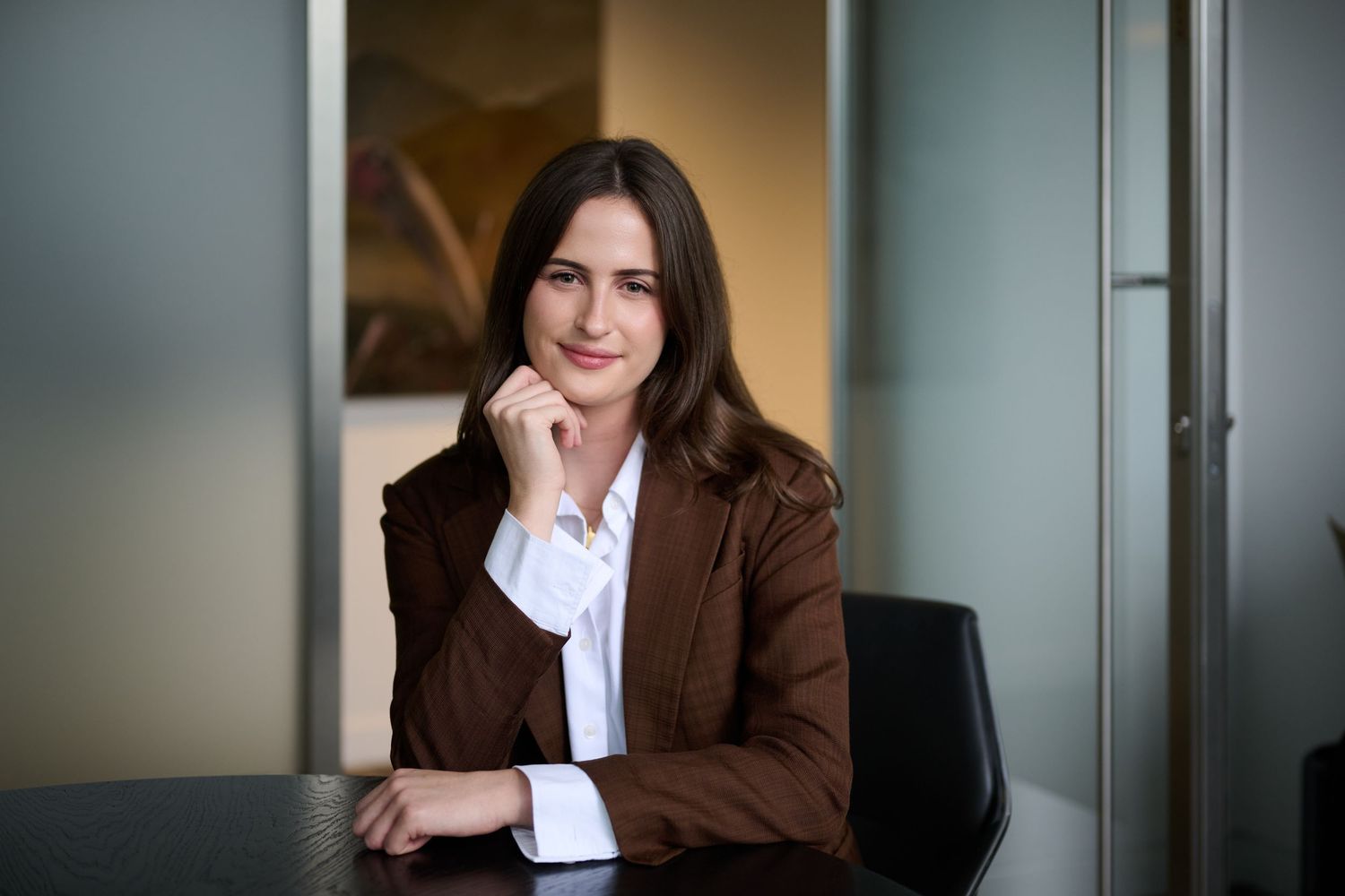 Professional woman in brown blazer sitting at desk with hand on chin in modern office setting.