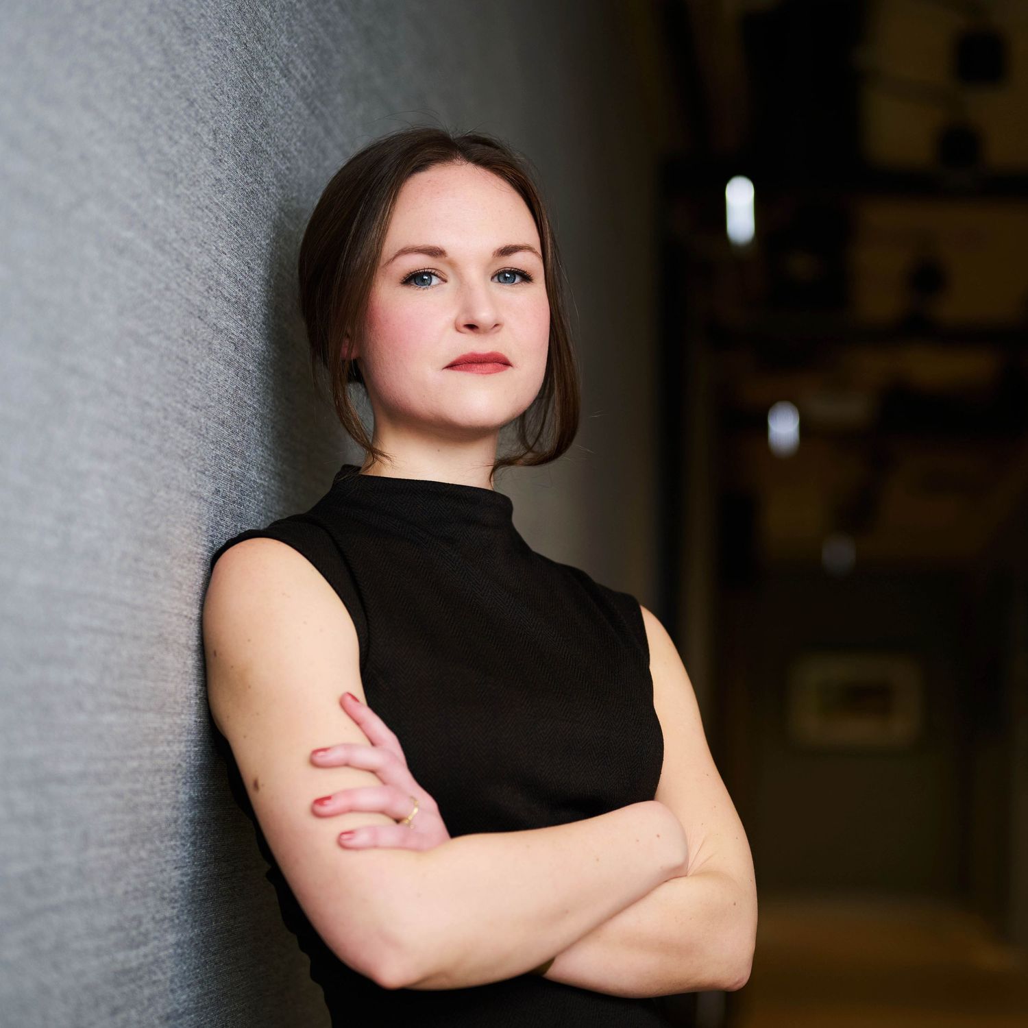 Young woman in black sleeveless top, arms crossed, leaning confidently against a dark textured wall.