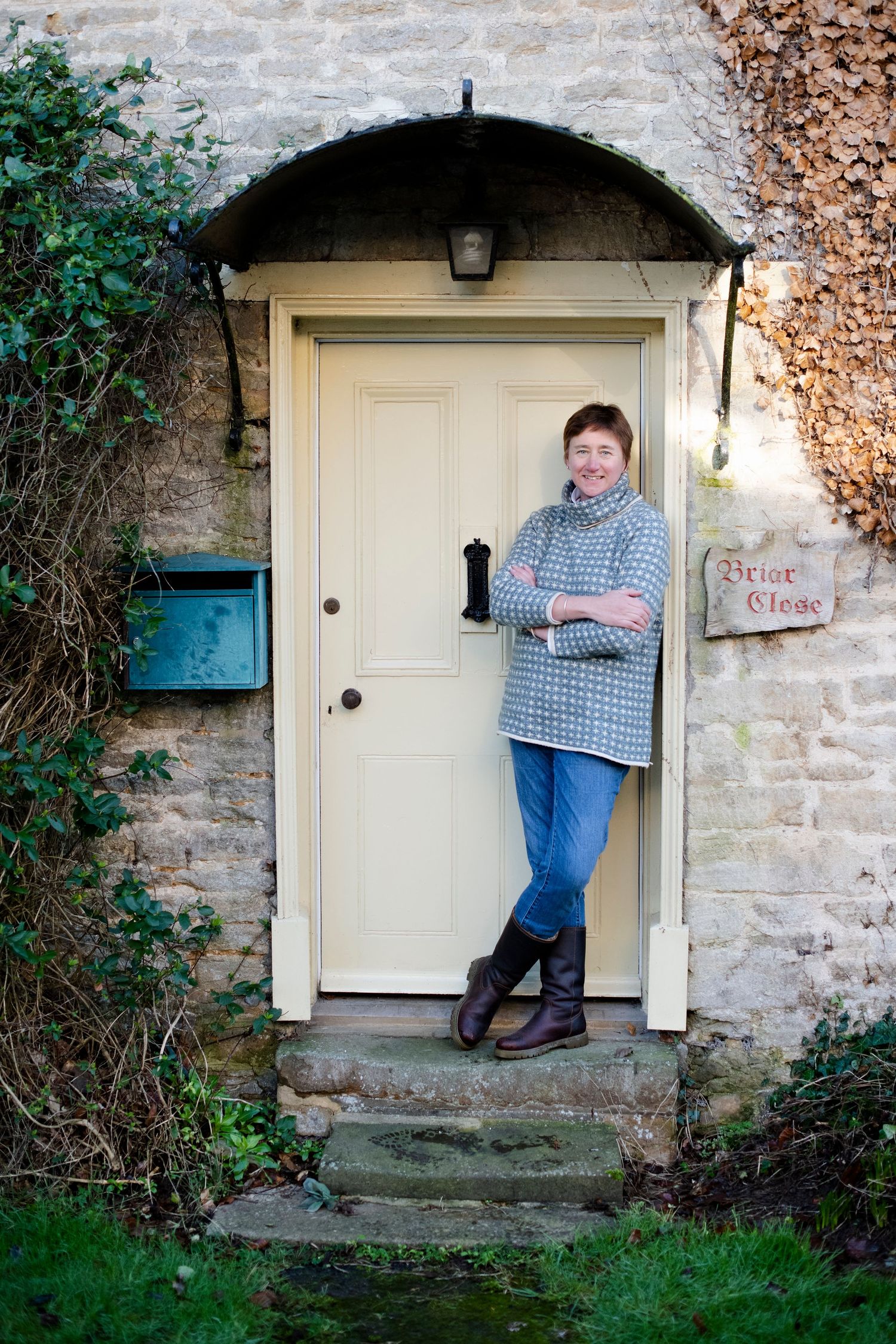 Woman in a houndstooth sweater and jeans leaning against a cream door of a charming stone cottage.