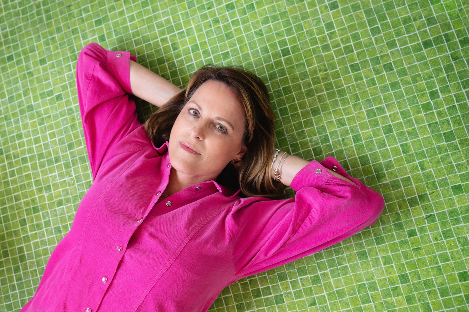 Woman in pink shirt lying on back on green mosaic tile floor looking up at camera.