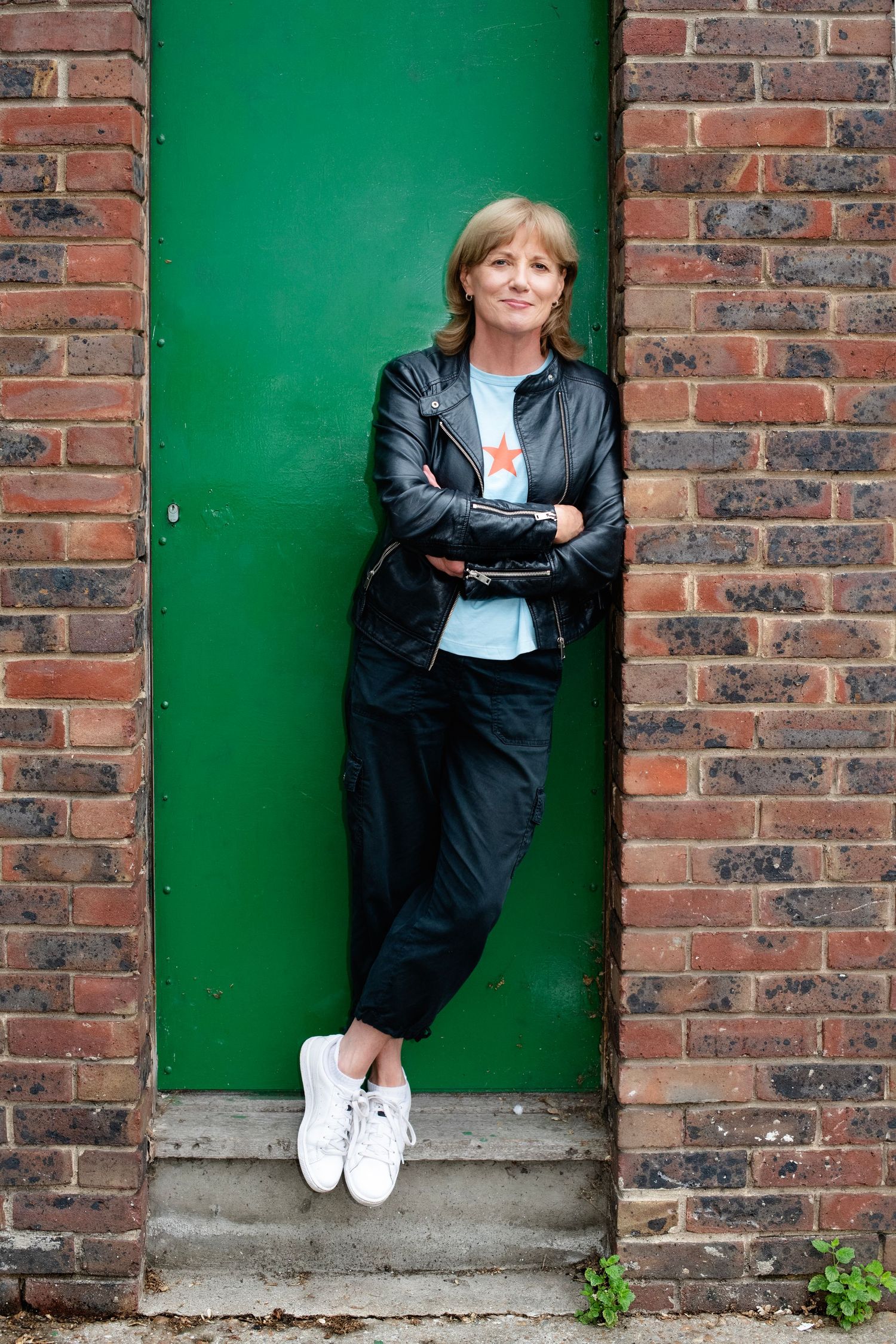 Blonde woman in black leather jacket and trousers leans against a green door framed by brick wall.